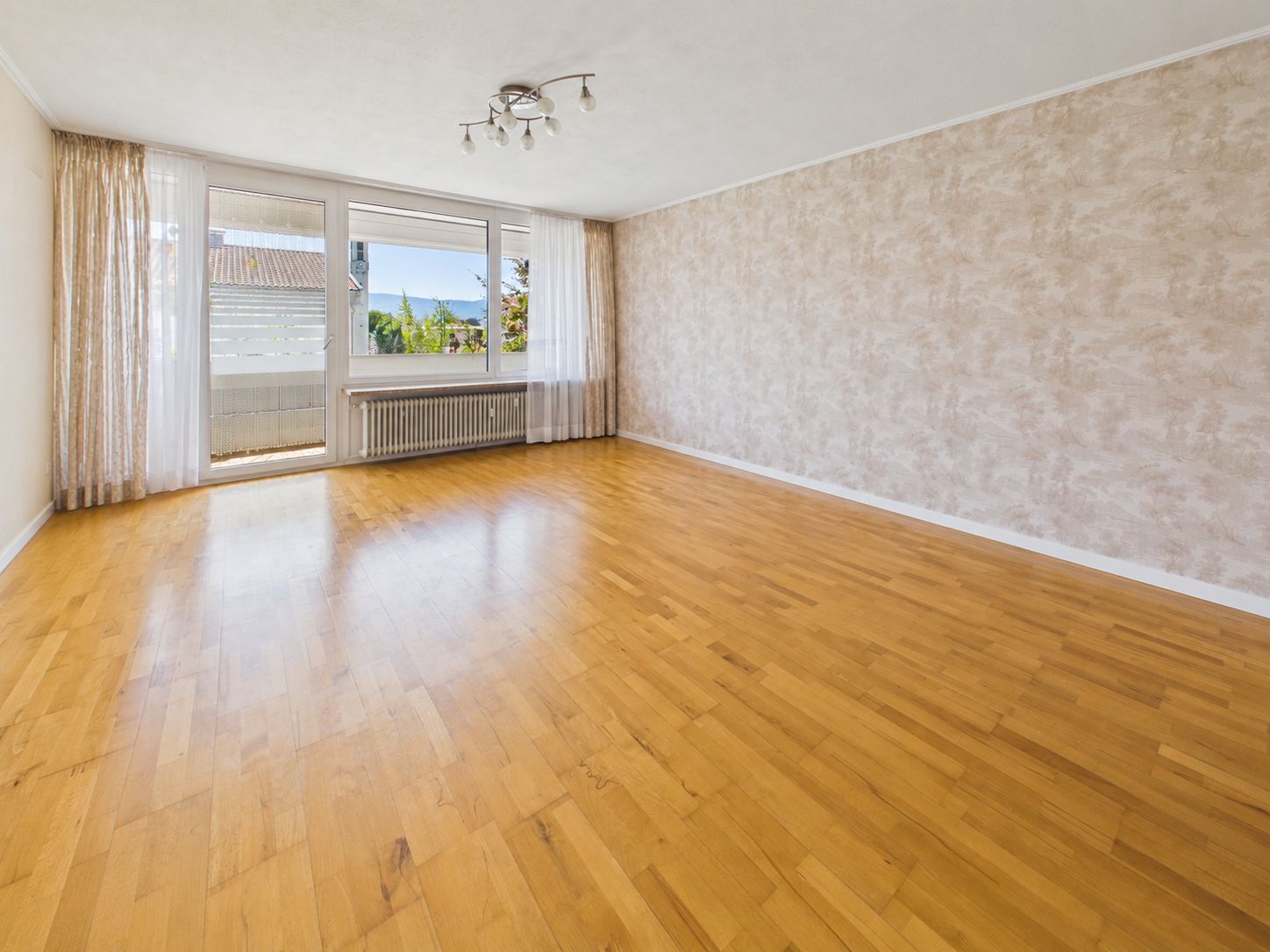 Helles, leeres Wohnzimmer mit Parkettboden, Fensterfront und Zugang zum Balkon.
