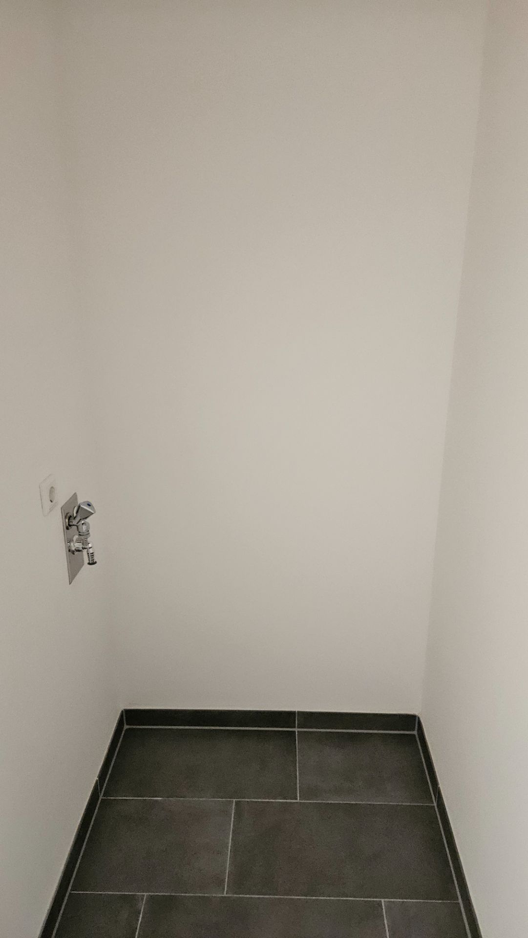 Kleiner leerer Raum mit weißen Wänden, dunklem Fliesenboden und einem Wasseranschluss.