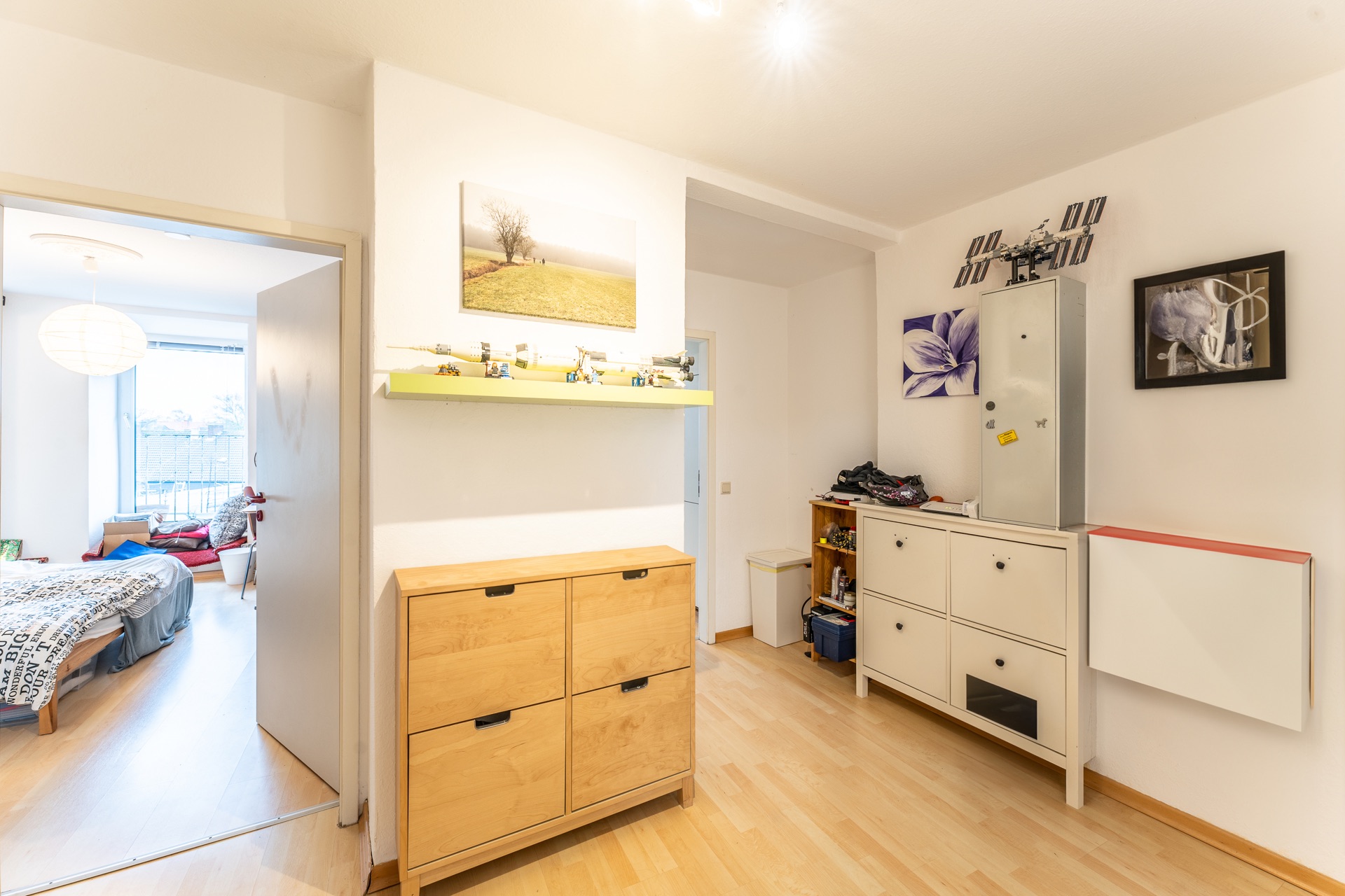 Heller Flur mit Holz- und weißen Schuhschränken, Wandbildern und Blick ins Schlafzimmer.