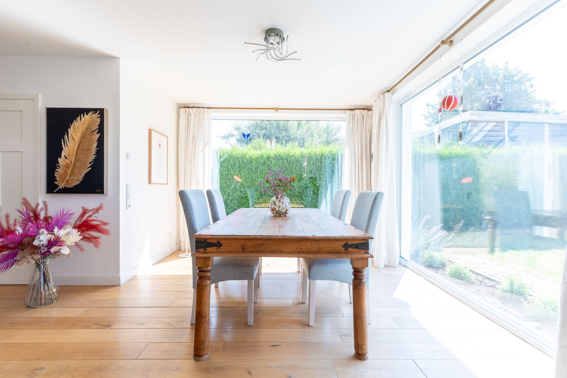 Esszimmer mit Holztisch, sechs Stühlen, Blumenstrauß und großen Fenstern mit Blick in den Garten.