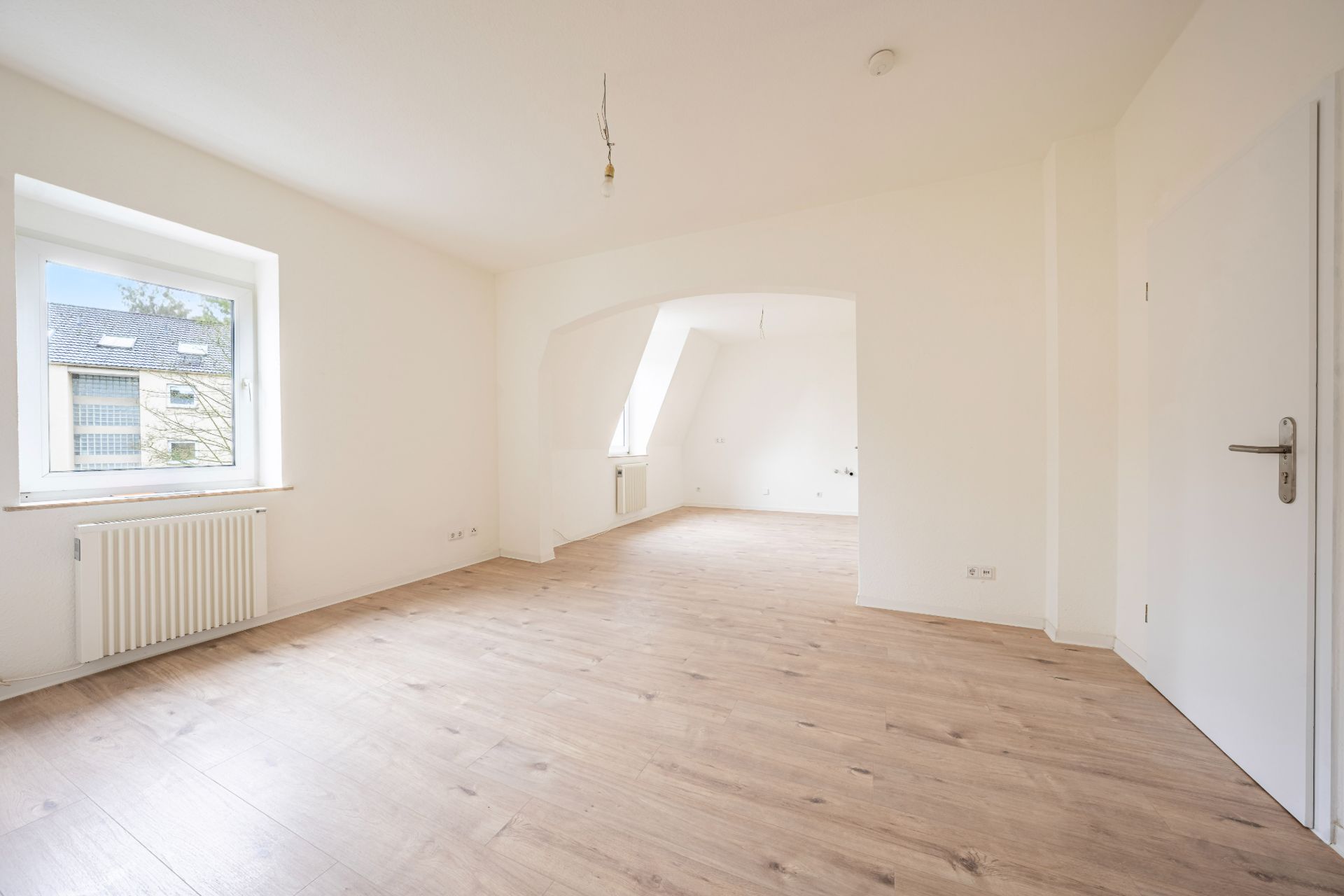 Helles, leeres Zimmer mit Holzfußboden, Fenster, Tür und weiß gestrichenen Wänden.