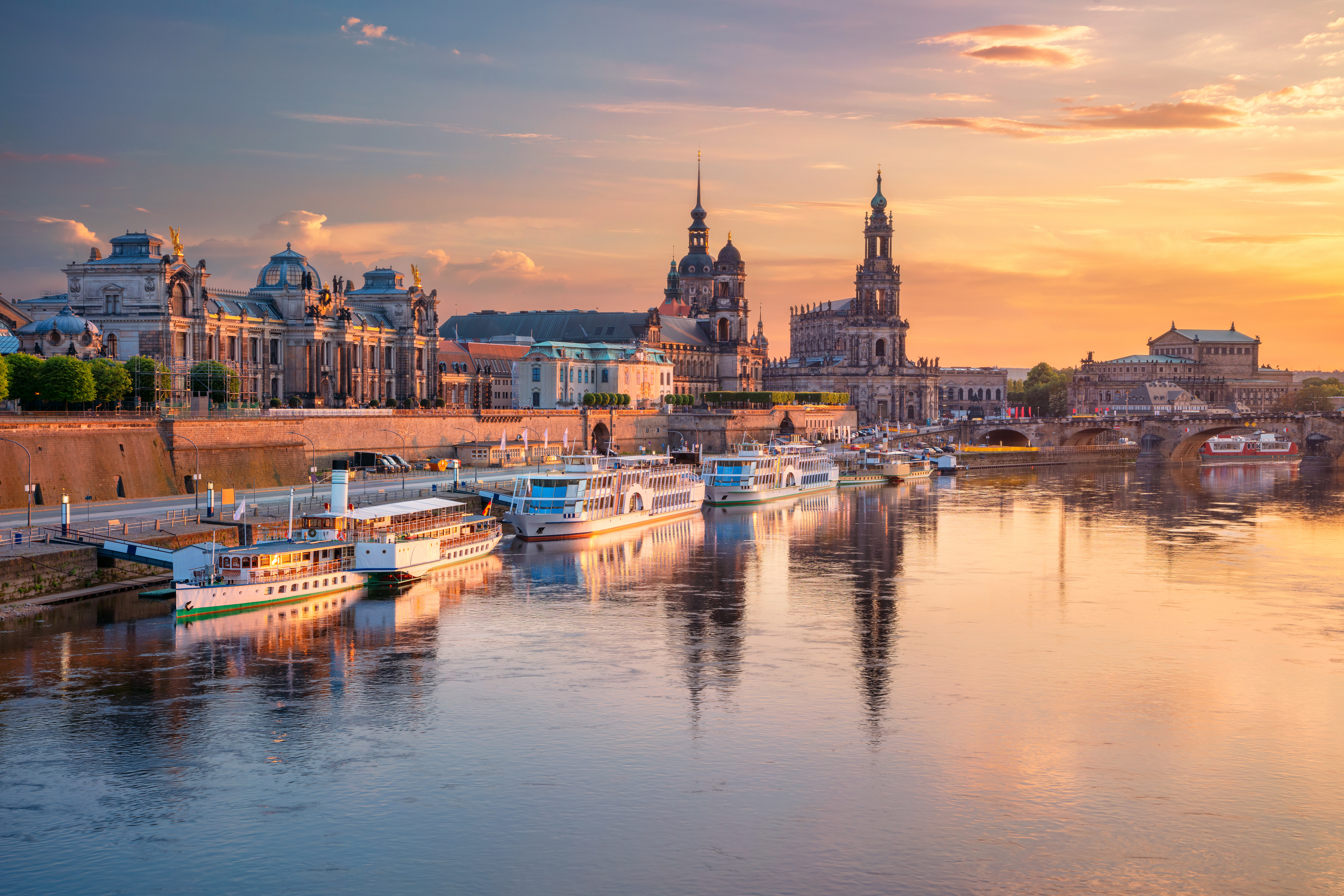 Fluss mit mehreren Booten und historischen Gebäuden im Hintergrund bei Sonnenuntergang.