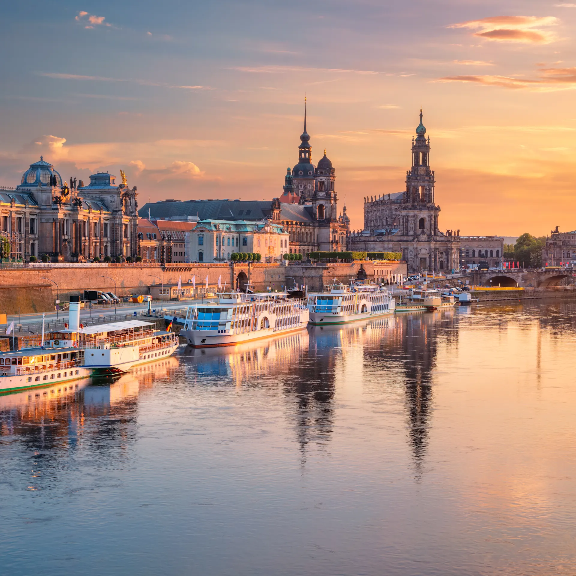 Fluss mit mehreren Booten und historischen Gebäuden im Hintergrund bei Sonnenuntergang.