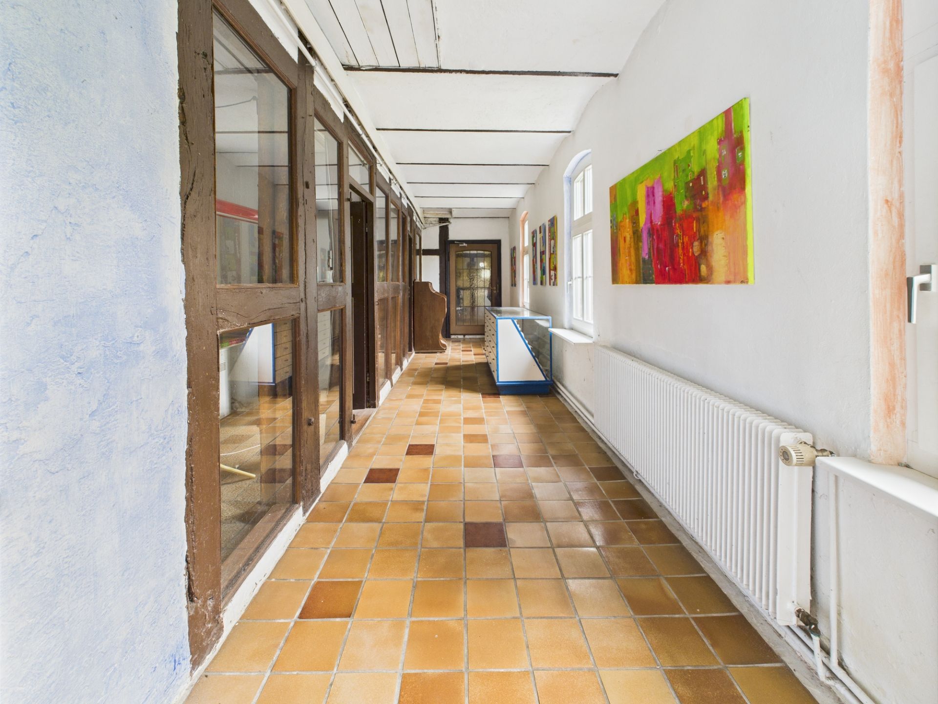 Langer Flur mit Fliesenboden, Fenstern, Heizkörpern und bunten Bildern an der Wand.