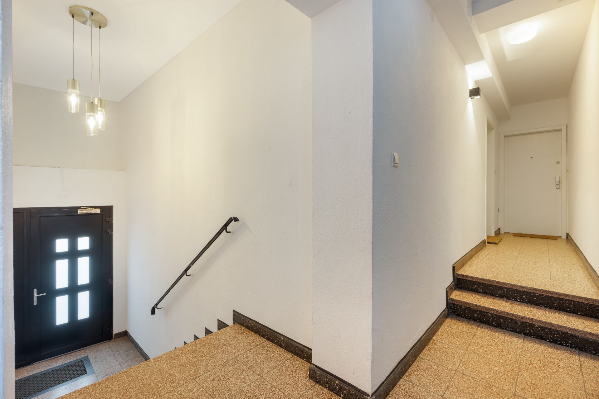 Hausflur mit Treppe, schwarzer Eingangstür und Flur zu Wohnungstüren, helle Wände und Deckenleuchte.