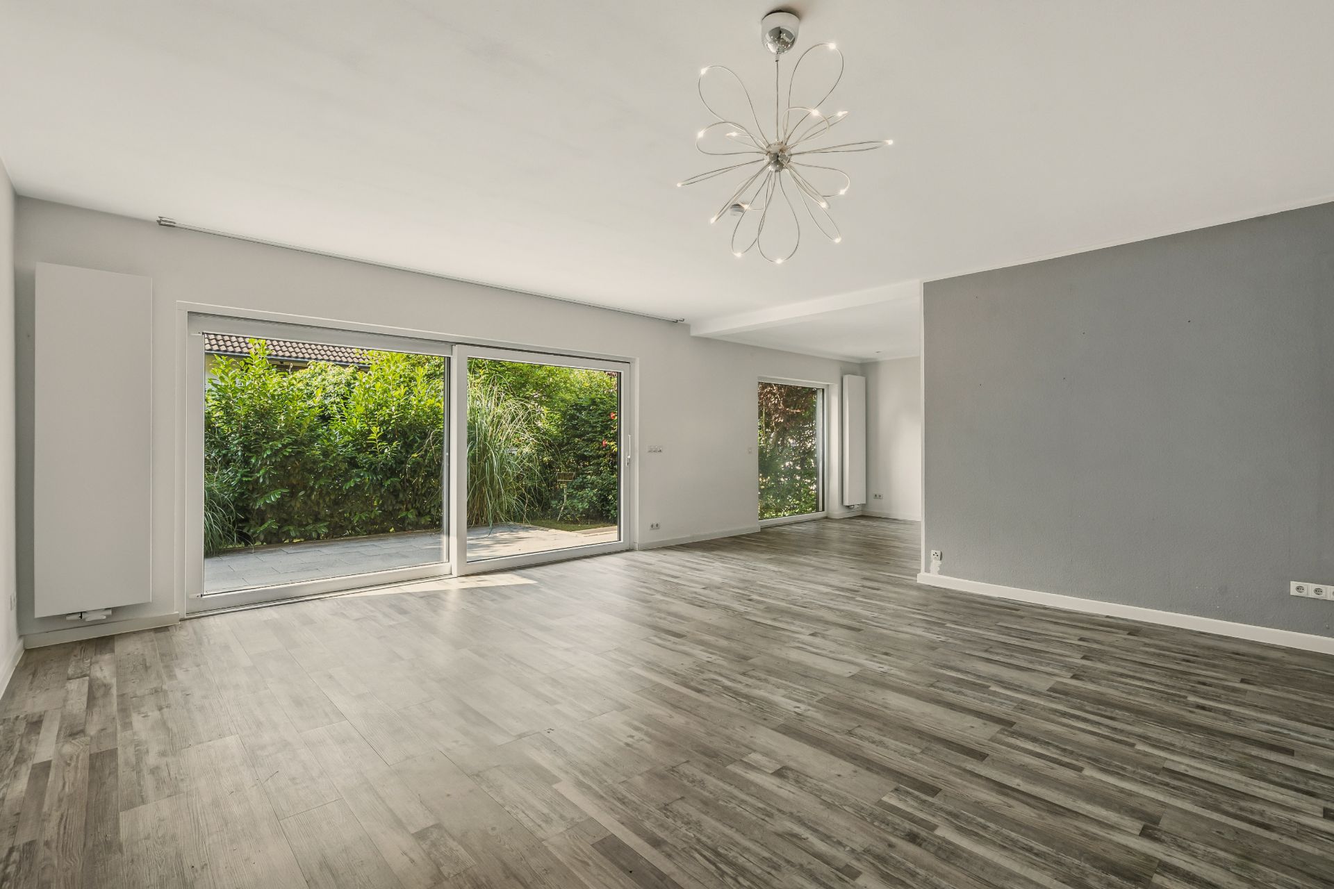 Helles, leeres Wohnzimmer mit großem Fenster, Glastür und Blick in den Garten.