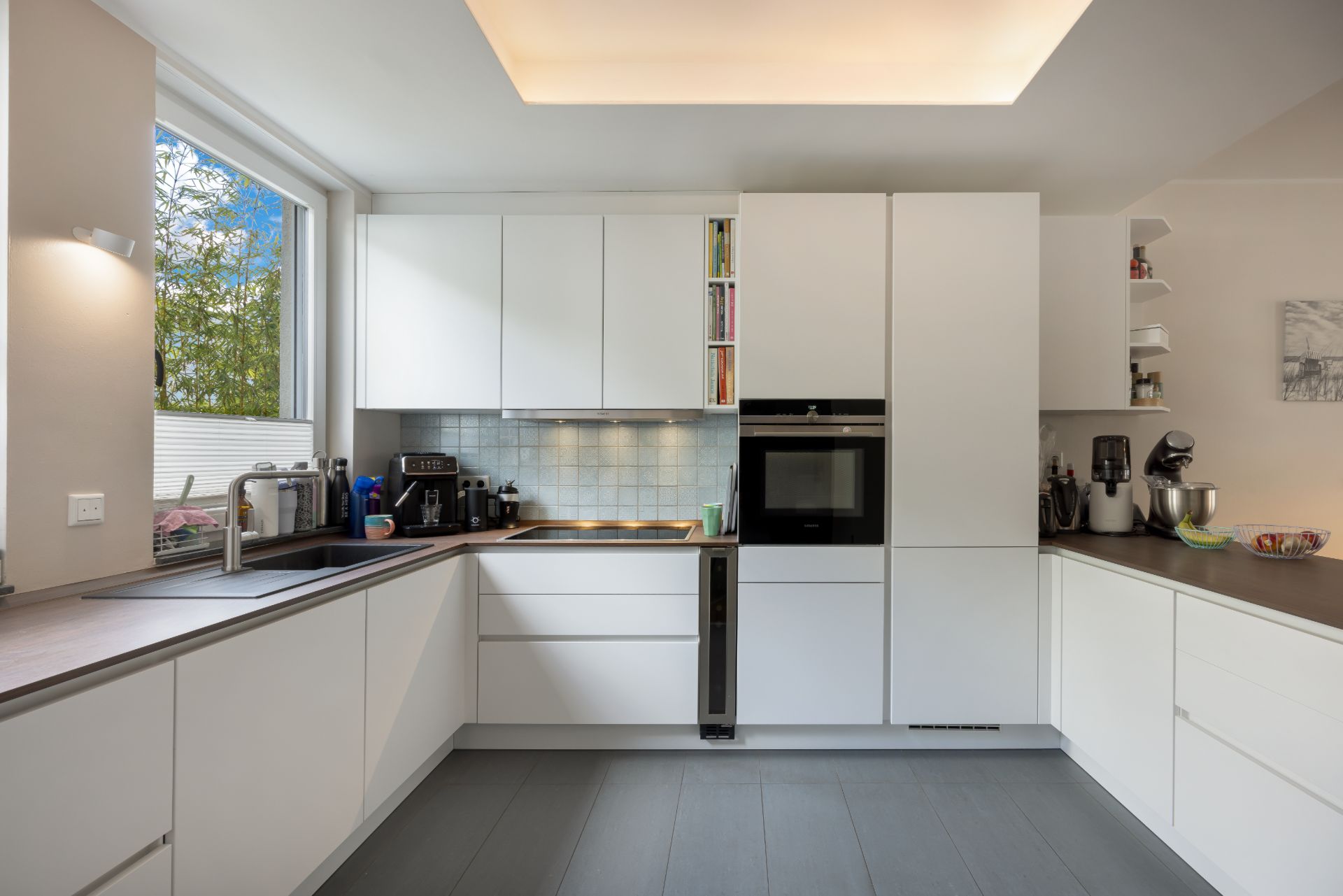 Moderne weiße Einbauküche mit großen Fenstern und Küchengeräten auf der Arbeitsplatte.