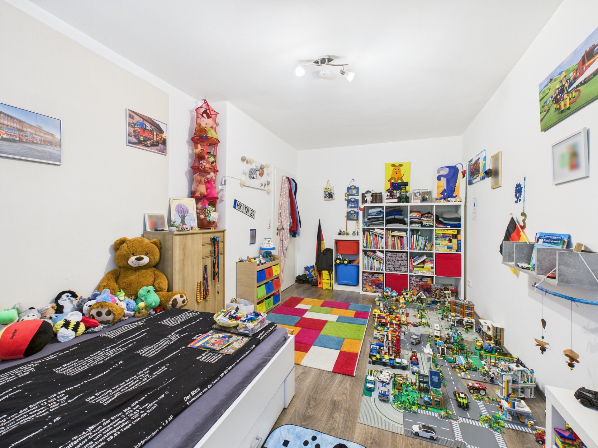 Kinderzimmer mit buntem Teppich, Spielzeug, Bett und Regalen voller Bücher und Spielsachen.