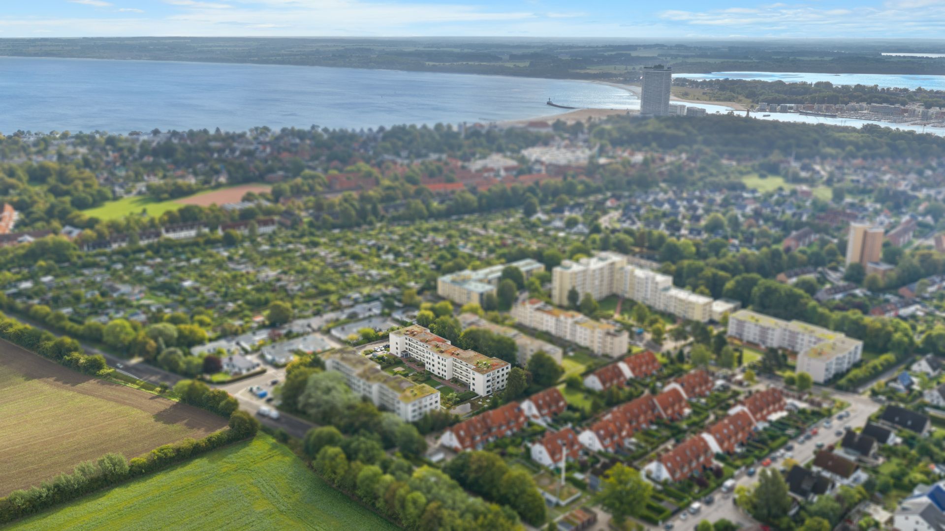 Luftaufnahme einer Wohnsiedlung mit mehreren Häusern neben Feldern und Wasser im Hintergrund.