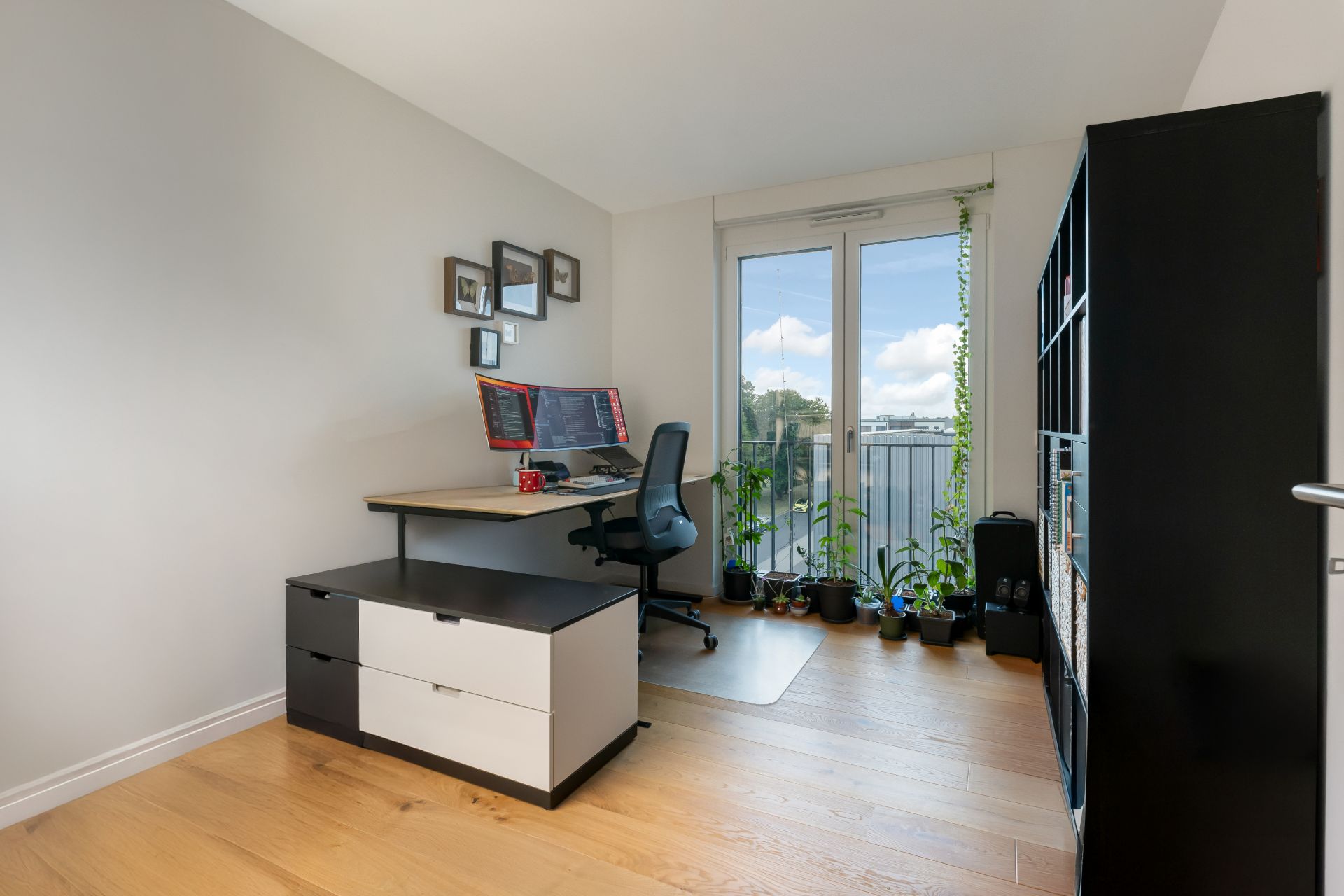 Modernes Arbeitszimmer mit Schreibtisch, Bürostuhl, Regal und Pflanzen vor einem Fenster.