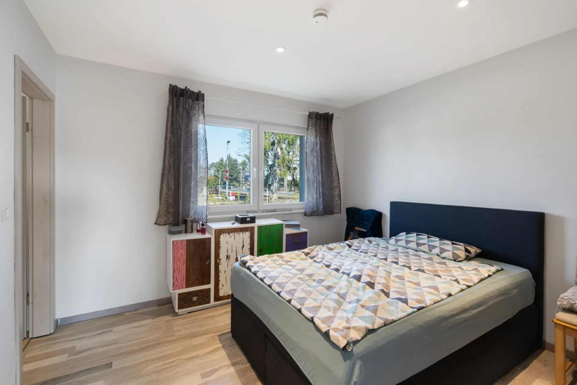 Modernes Schlafzimmer mit großem Bett, Kommode unter Fenster und Holzboden.