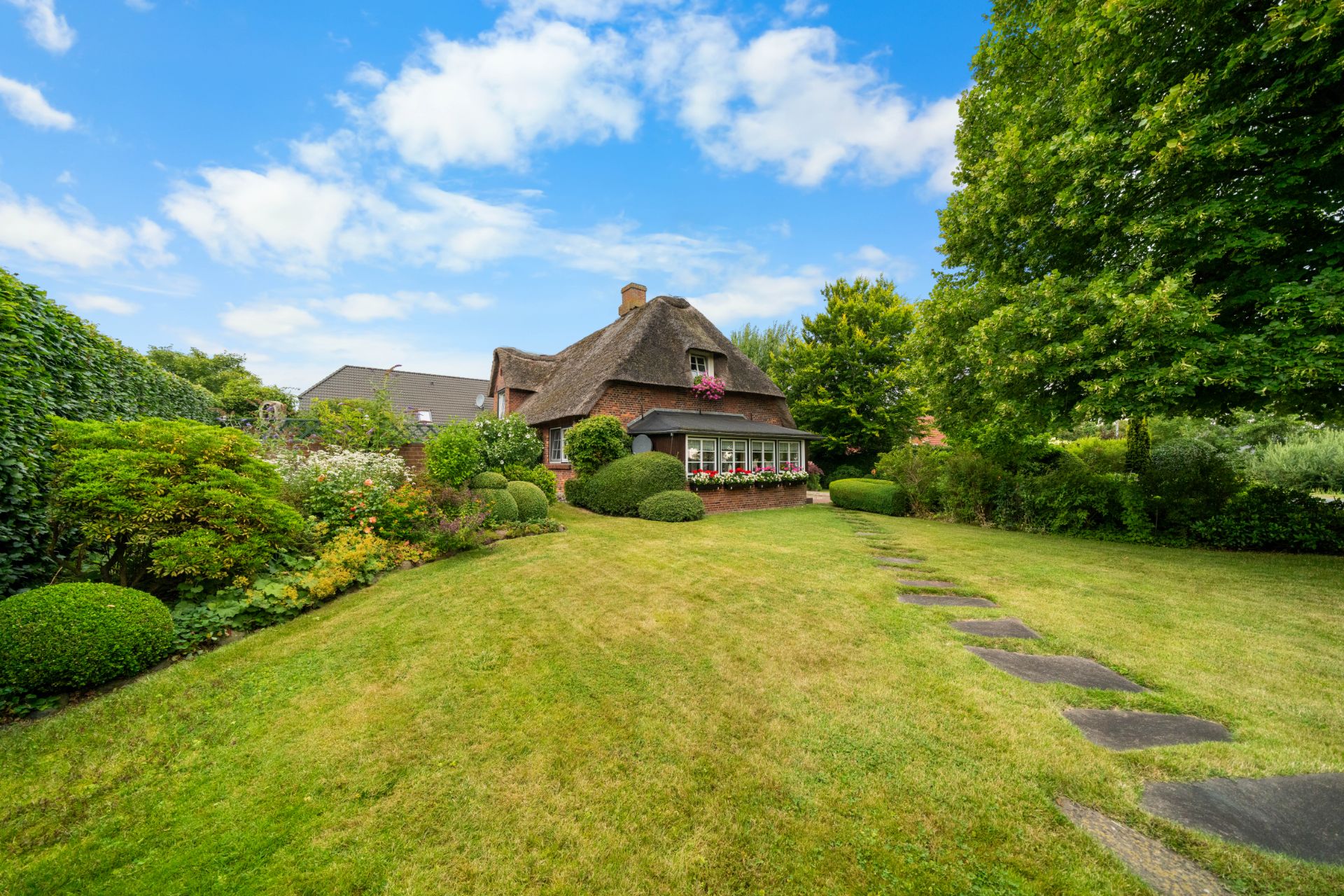Haus mit Reetdach und großem gepflegtem Garten, umgeben von Bäumen und Sträuchern.