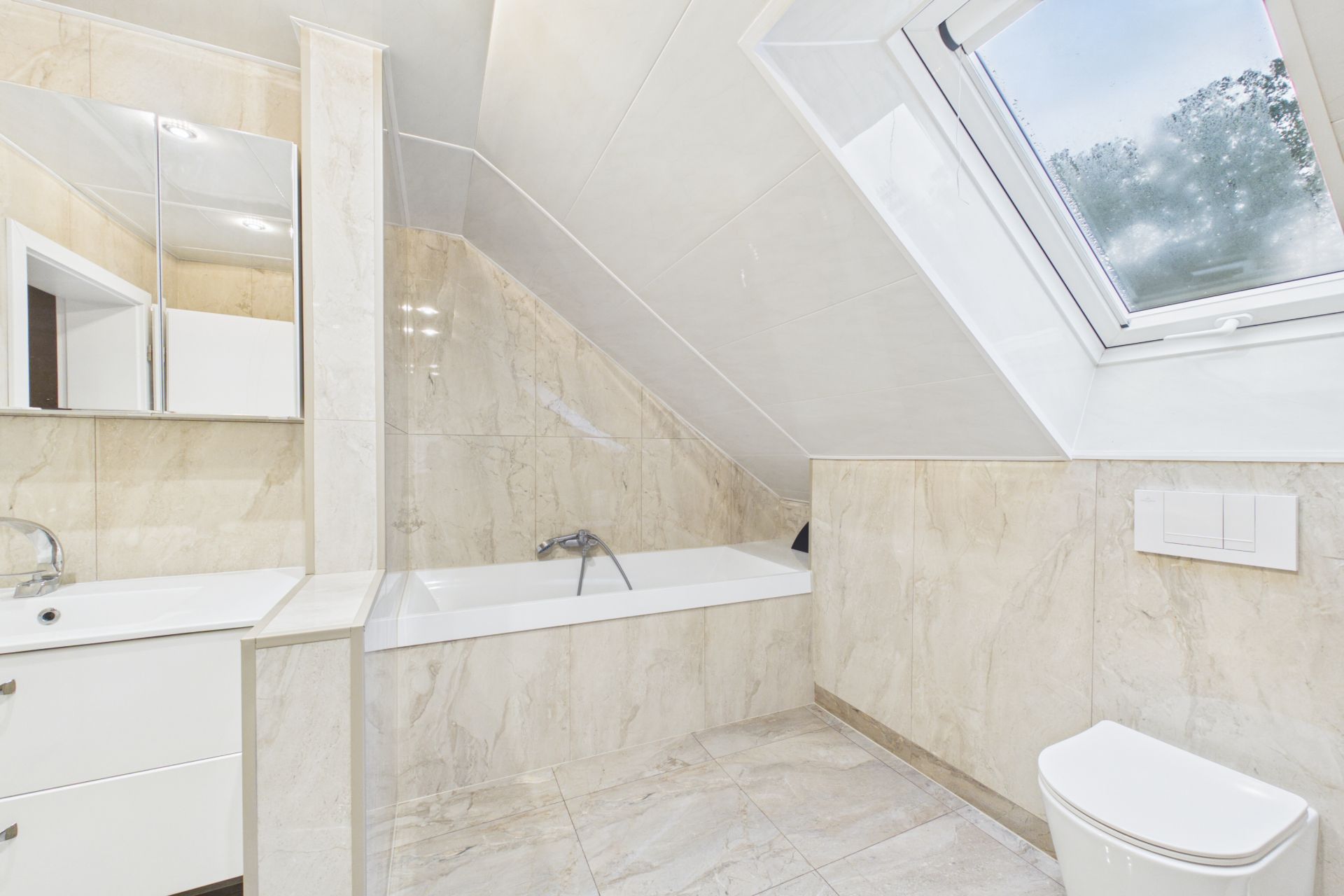 Helles Badezimmer mit Badewanne, Waschbecken, Spiegel und Fenster im Dachgeschoss.