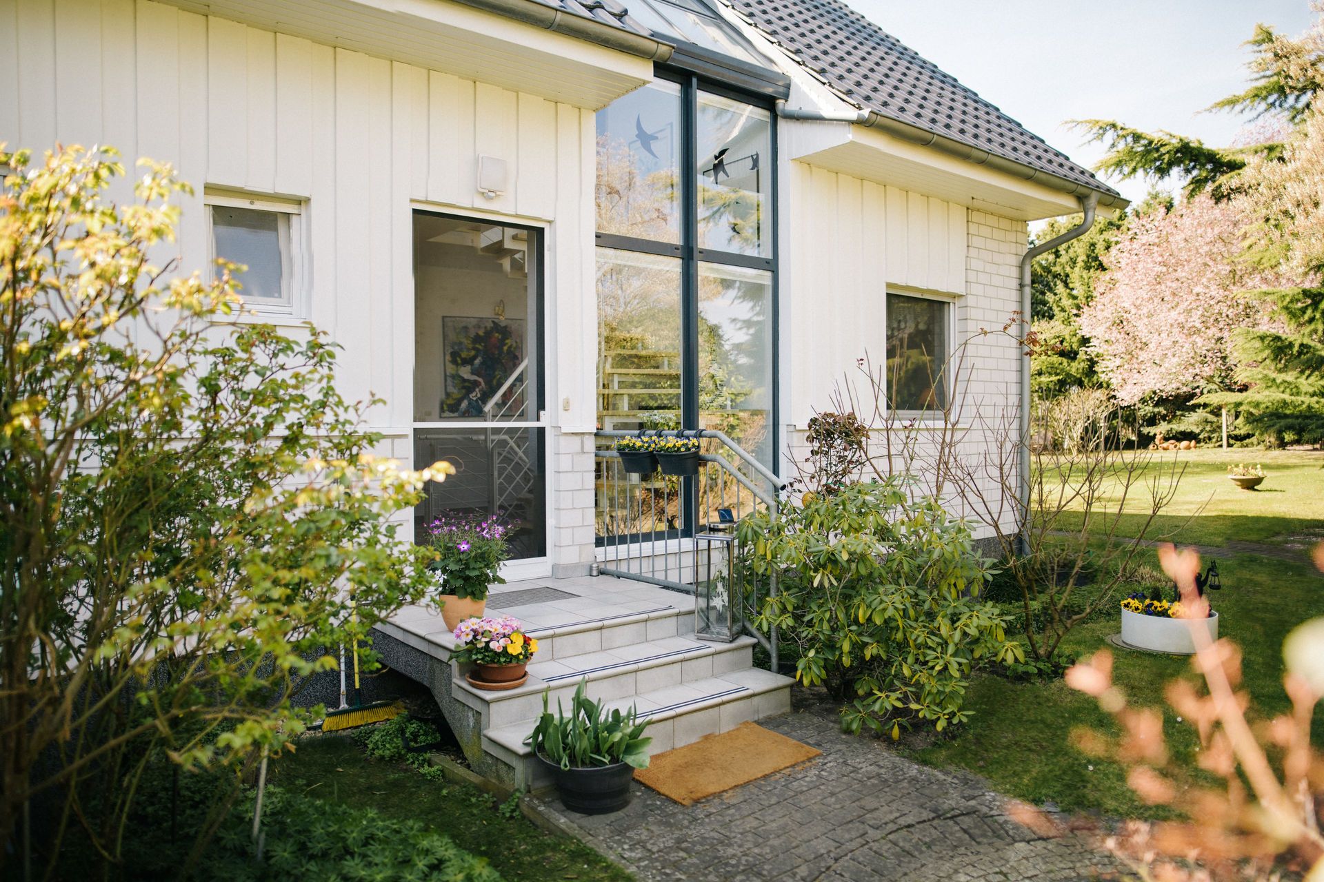 Eingang eines weißen Hauses mit Treppe, Pflanzen und Garten im Vordergrund.