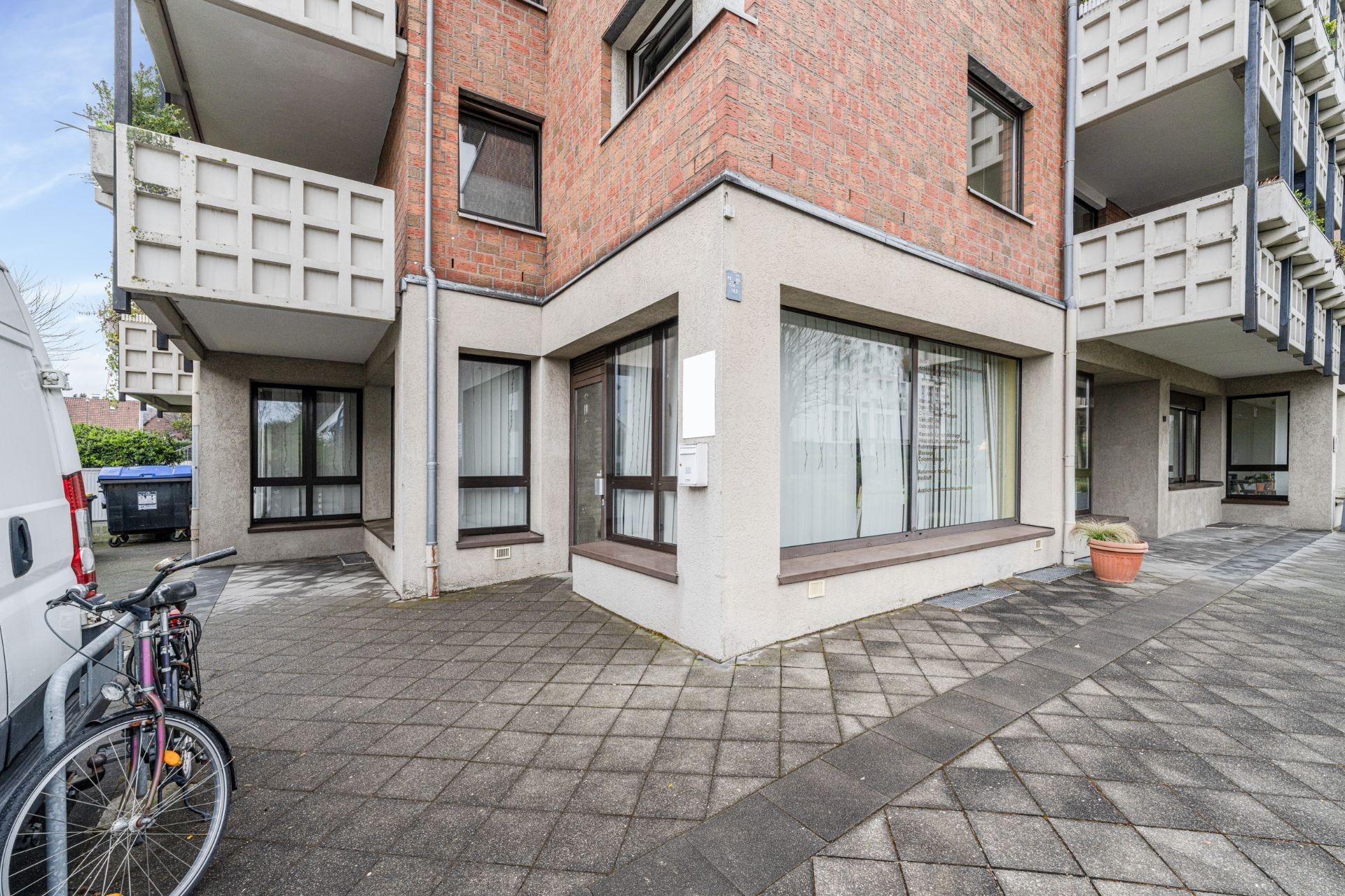 Eckwohnung mit großen Fenstern, Balkon, Fahrradständer und Blumenkübel auf gepflastertem Gehweg.