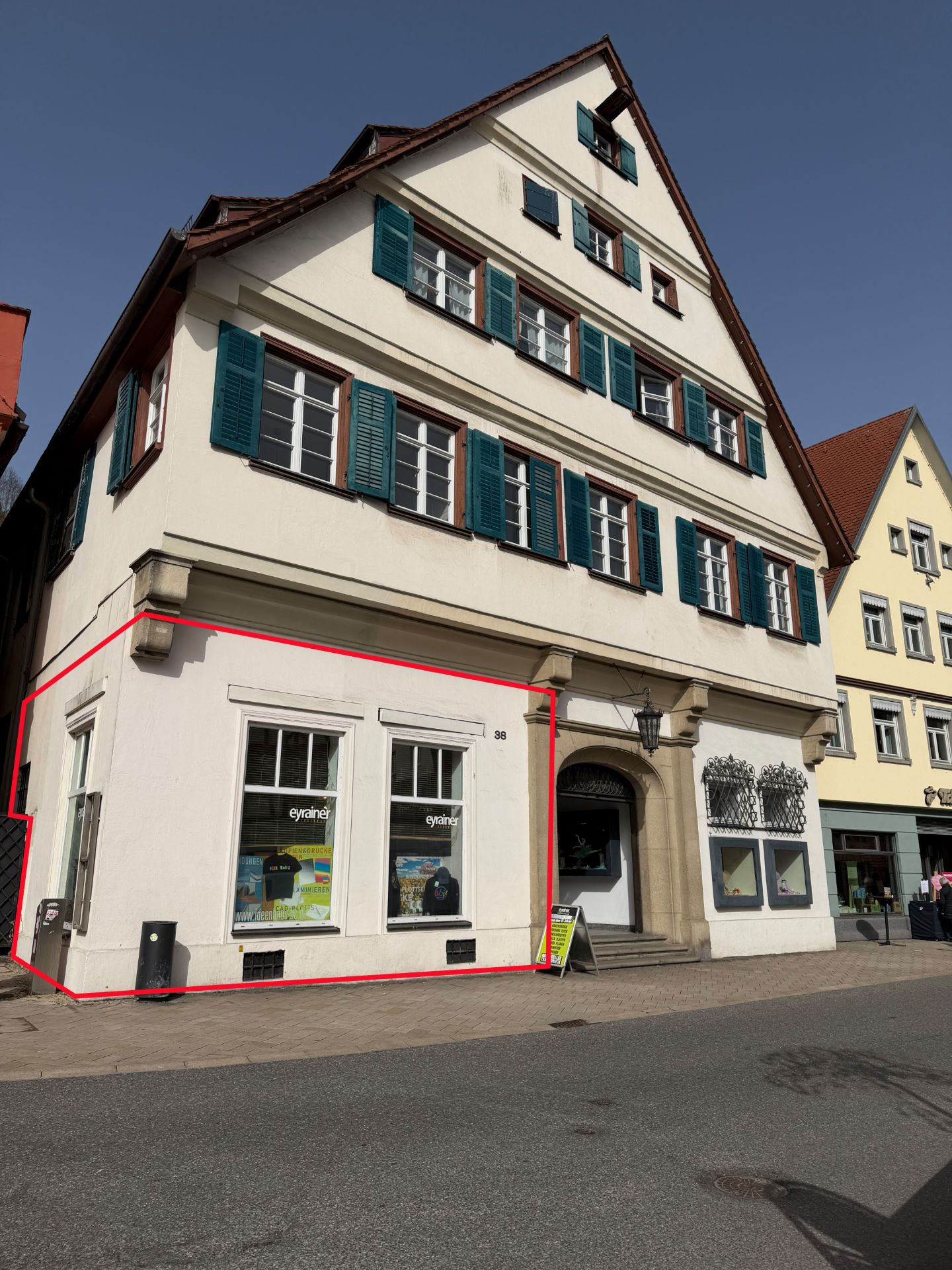 Wohnhaus mit Geschäft im Erdgeschoss, weiße Fassade mit vielen Fenstern und grünen Fensterläden.