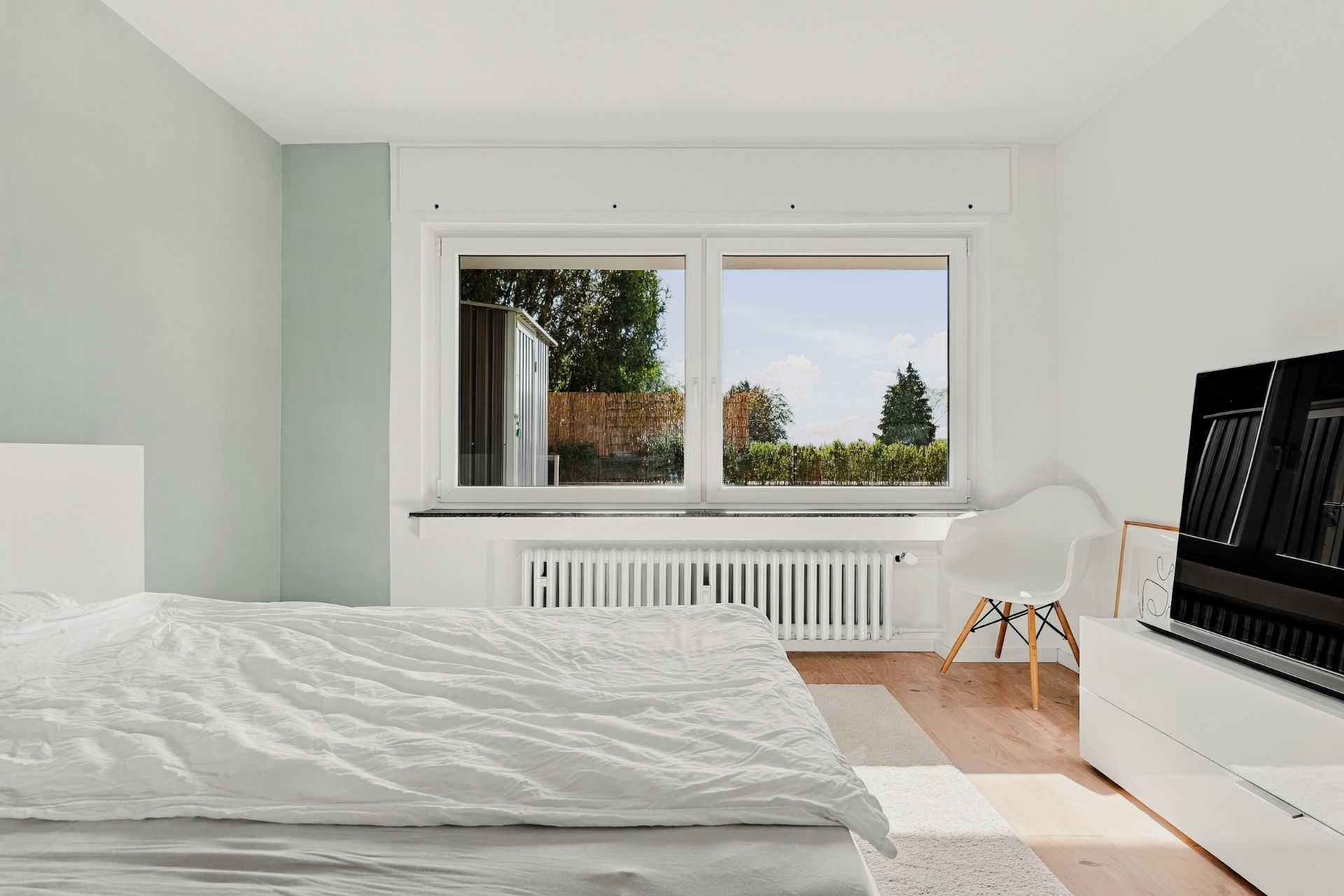 Helles Schlafzimmer mit Bett, großem Fenster, Stuhl und Kommode mit Fernseher.
