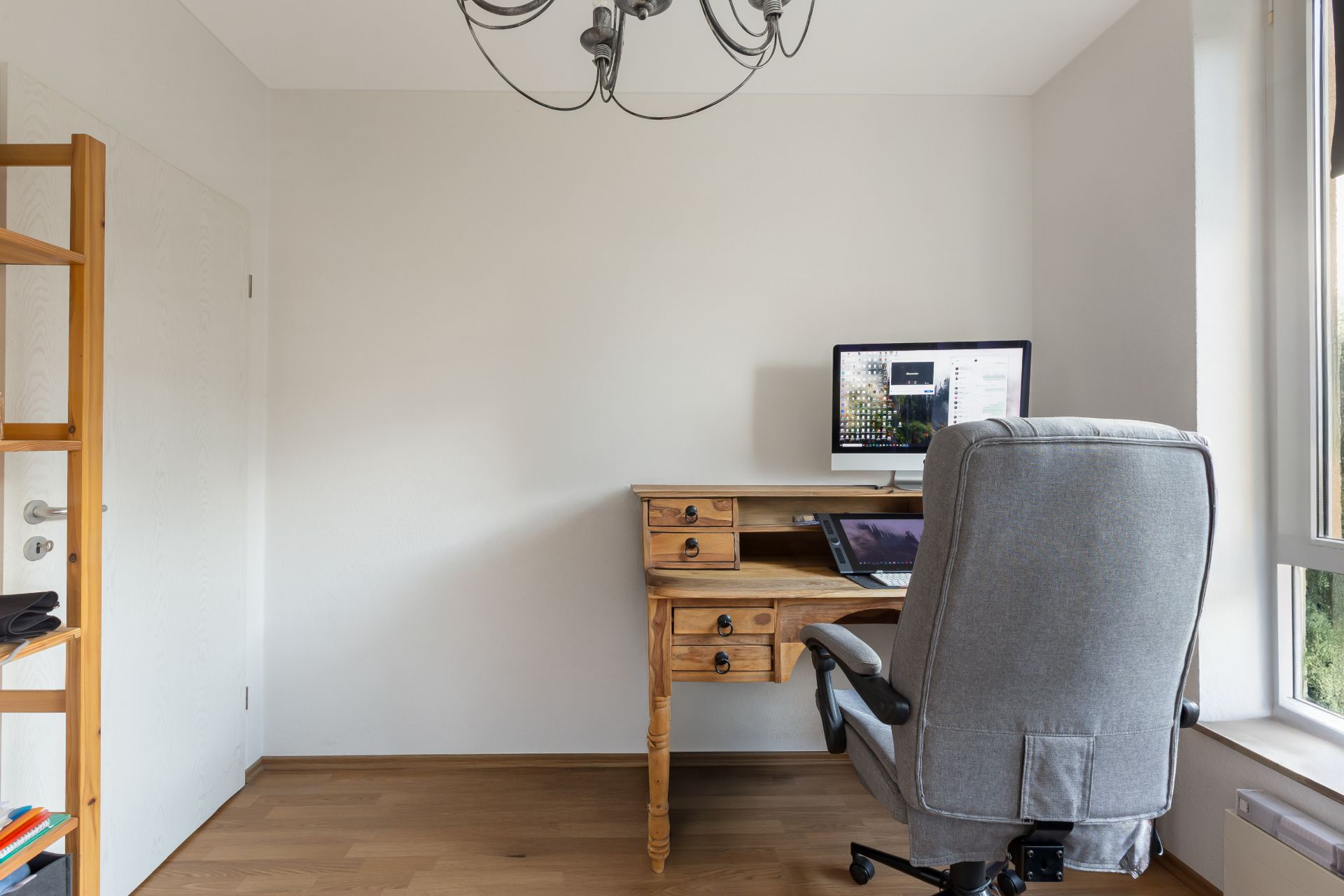 Kleines Arbeitszimmer mit Schreibtisch, Stuhl, Computer und Regal, Fenster rechts.