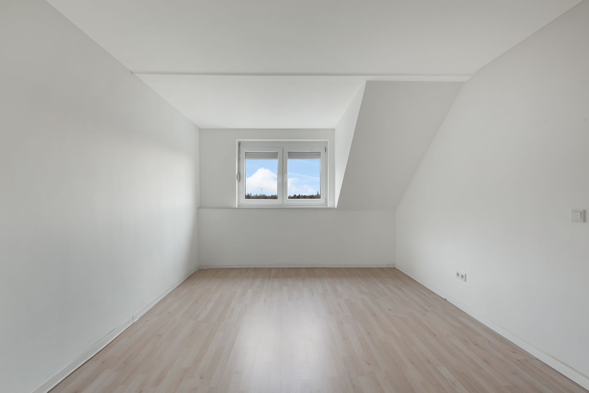 Leerer, heller Raum mit weiß gestrichenen Wänden, Holzfußboden und Fenster mit Blick nach draußen.