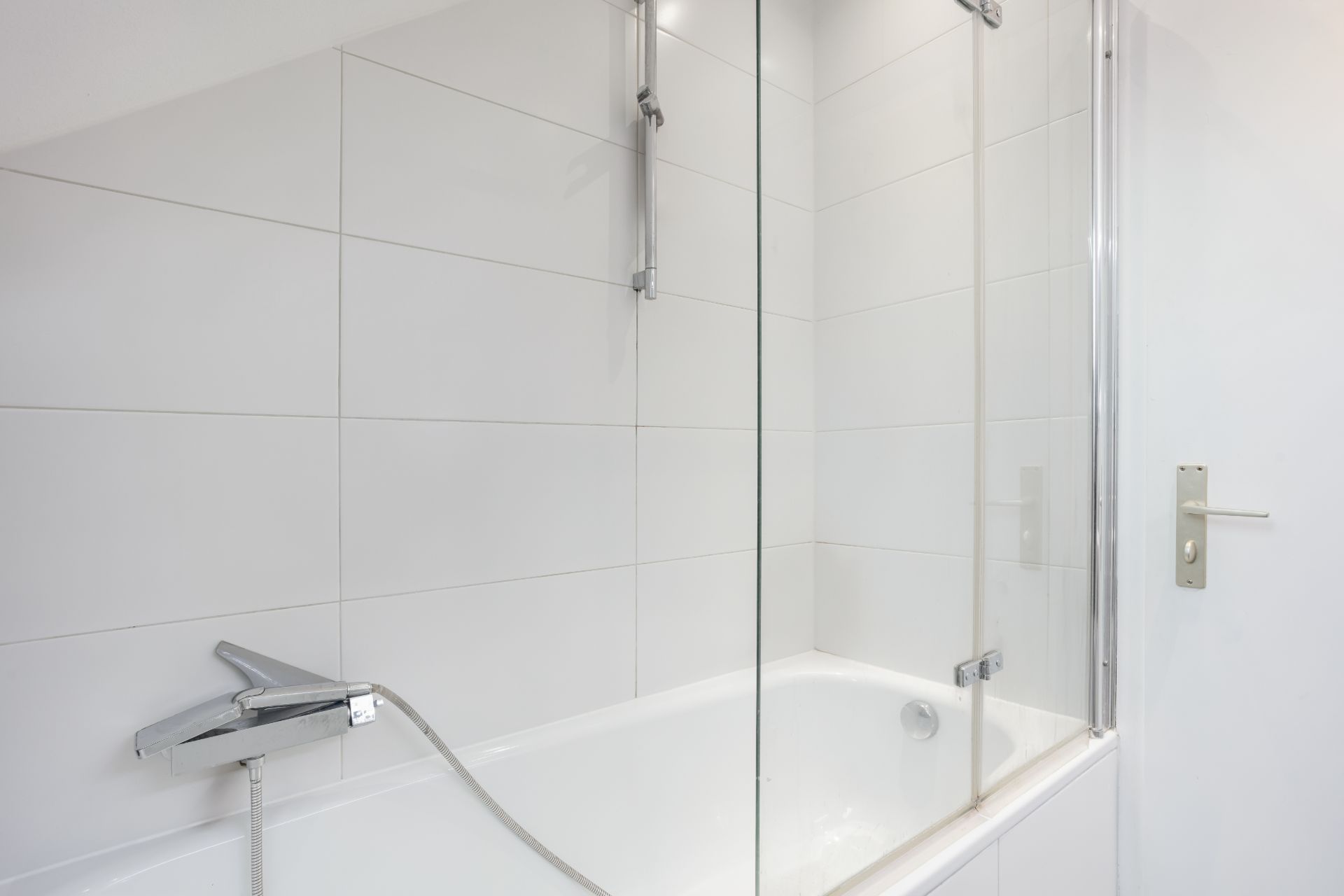 Weiße Badewanne mit Duscharmatur, Glasabtrennung und weißen Wandfliesen.
