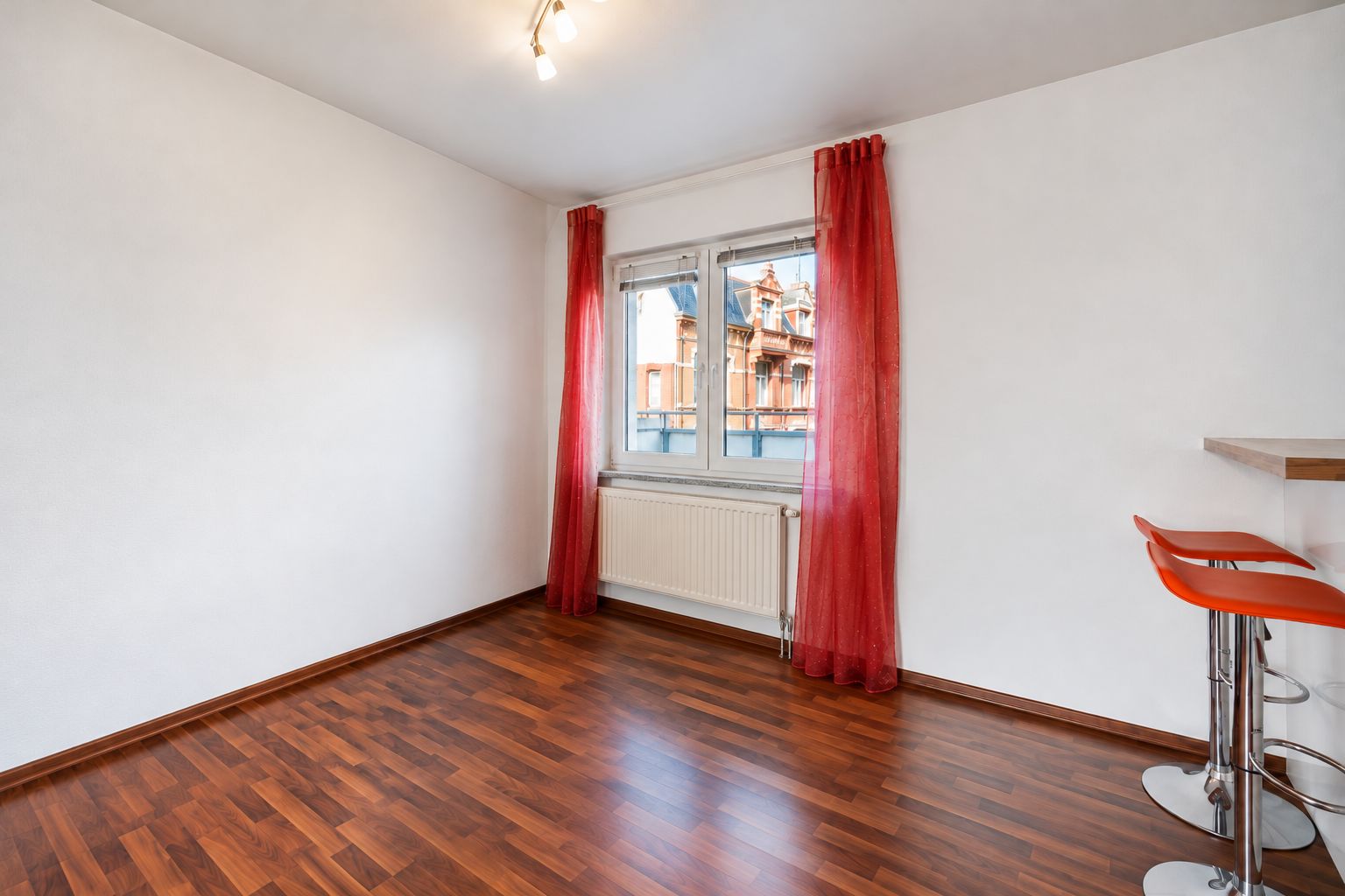 Helles Zimmer mit Holzfußboden, rotem Vorhang, Fenster und zwei Barhockern an einem Tresen.