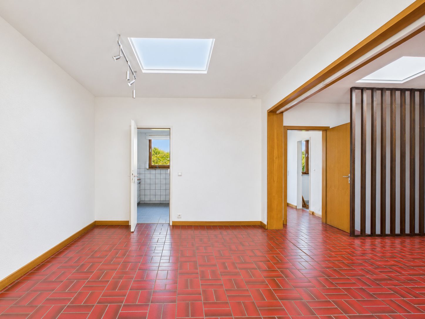 Heller Raum mit rotem Fliesenboden, weißen Wänden und Oberlichtern, Türen führen in andere Zimmer.