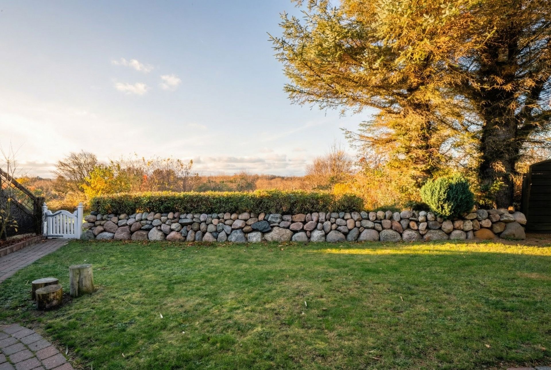 Garten mit Rasenfläche, Steinmauer, Bäumen und kleinem weißen Zaun.