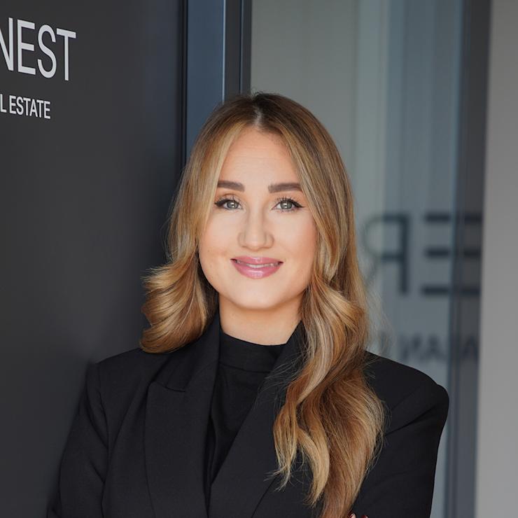 Frau mit langen, hellbraunen Haaren in schwarzem Blazer steht lächelnd vor einer dunklen Wand.