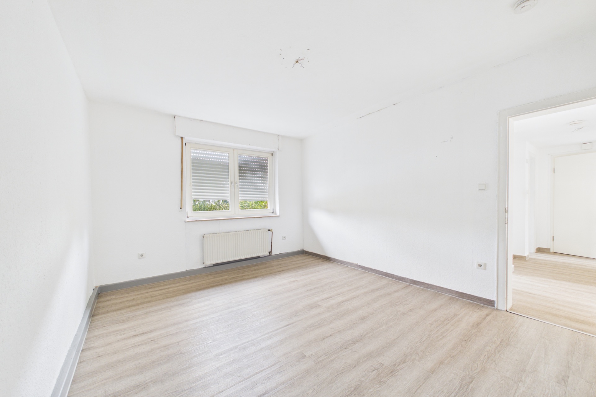 Leerer, heller Raum mit Holzfußboden, weißer Wand und Fenster mit Blick ins Grüne.