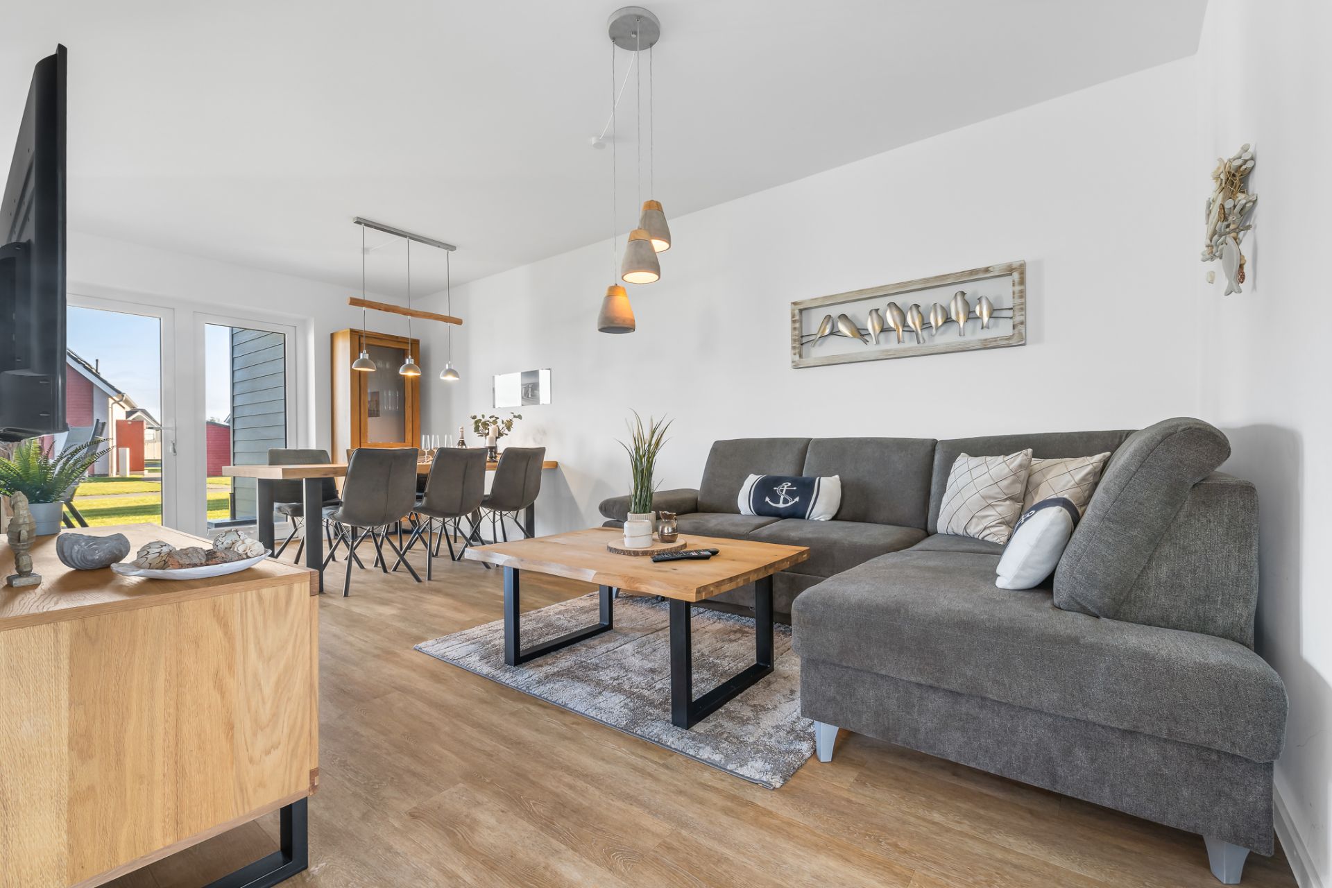 Modernes Wohnzimmer mit grauem Sofa, Holztisch und Essbereich neben großen Fenstern.
