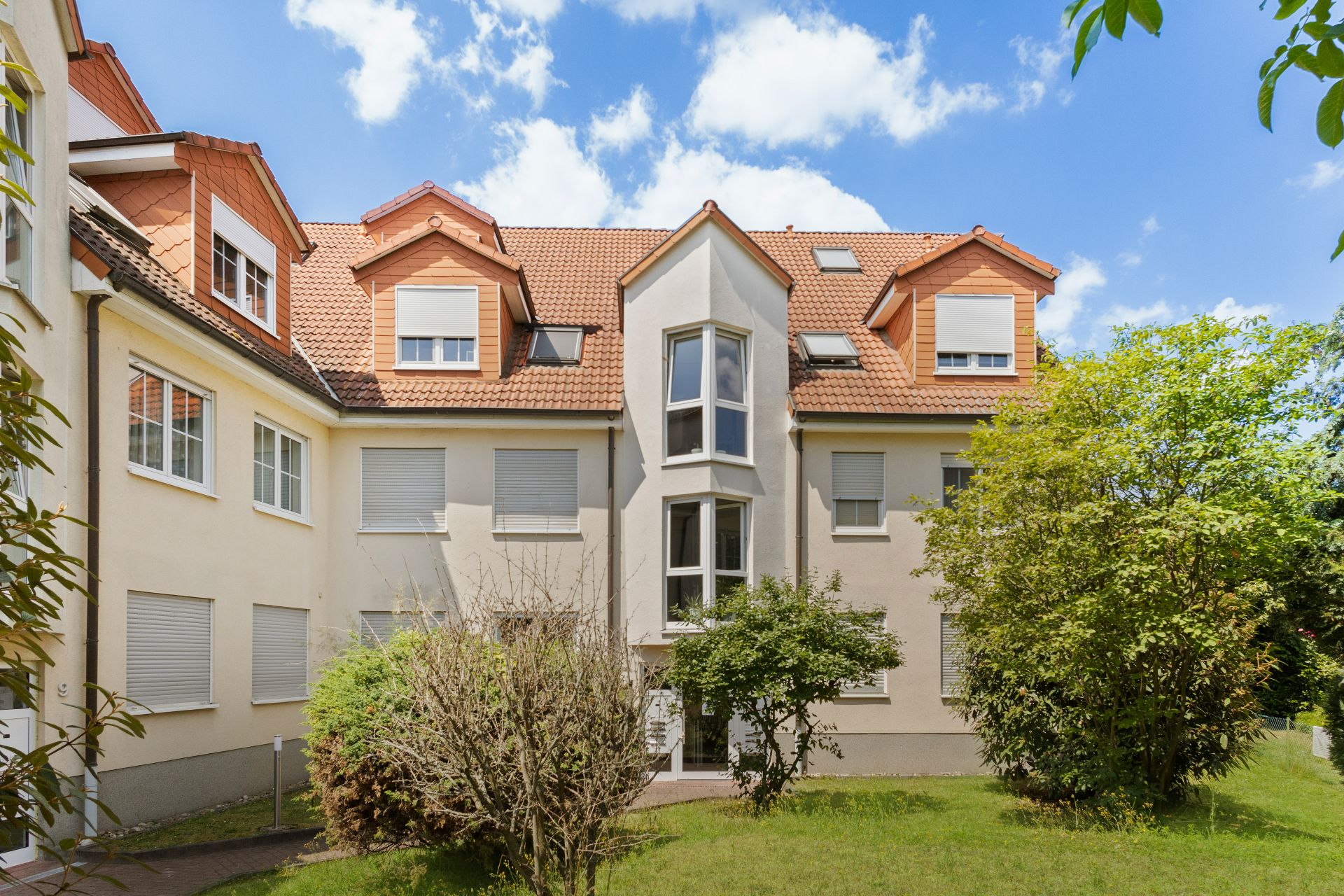 Mehrfamilienhaus mit roten Dachziegeln, mehreren Fenstern und Garten im Vordergrund.