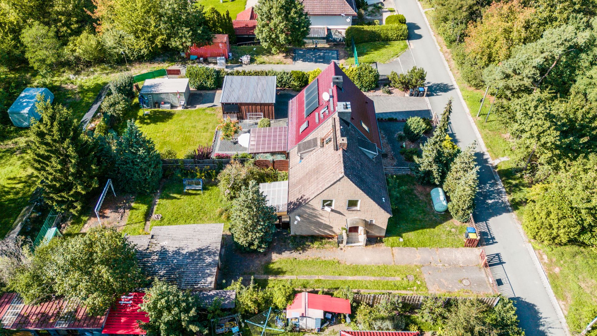 Luftaufnahme eines Wohnhauses mit Garten, Bäumen und Nebengebäuden an einer Straße.