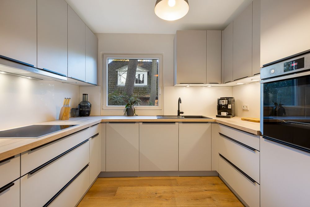 Moderne Einbauküche mit hellen Schränken, Herd, Spüle, Kaffeemaschine und Fenster.