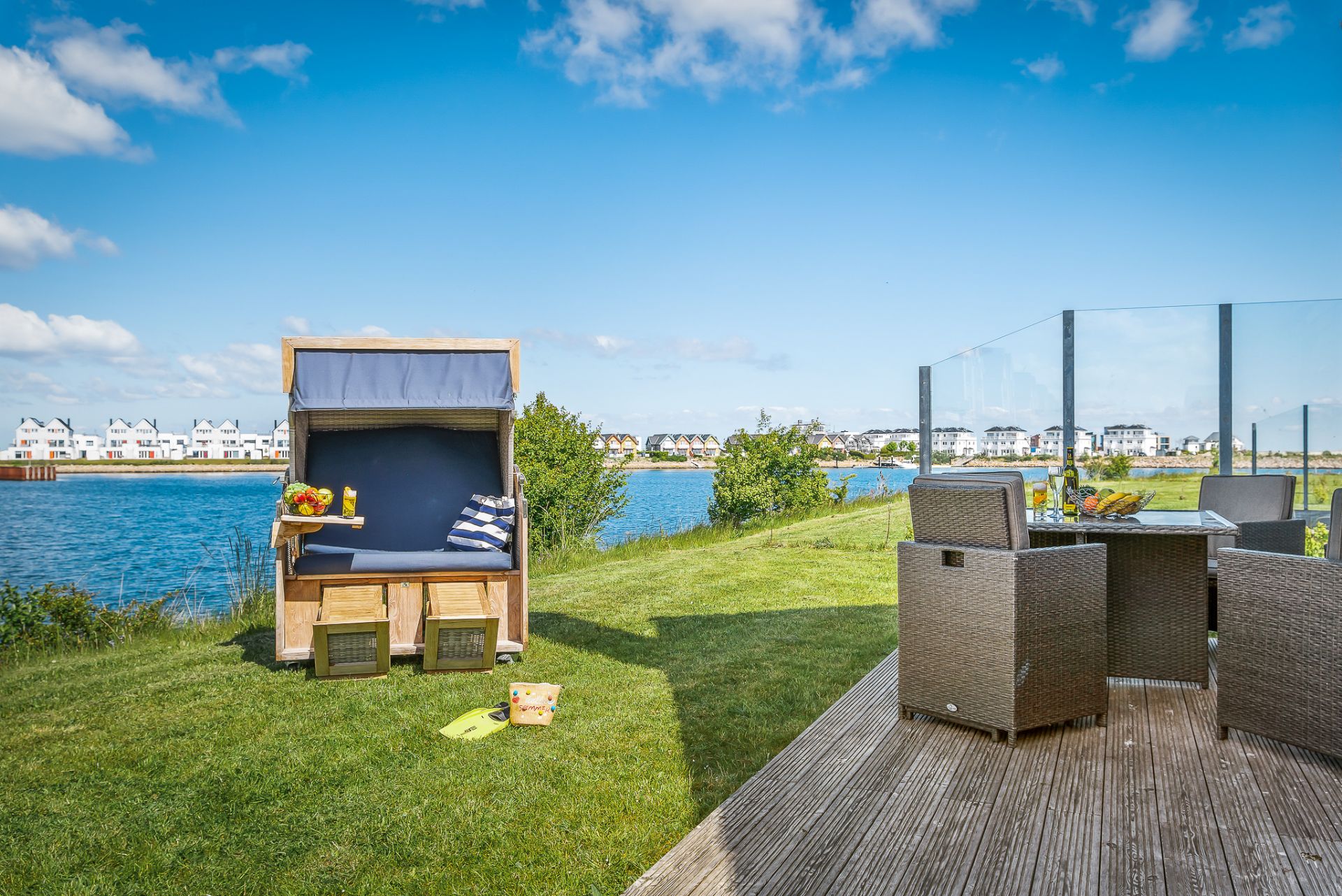 Garten mit Strandkorb, Holztisch und Stühlen, Blick auf einen See und Häuser im Hintergrund.
