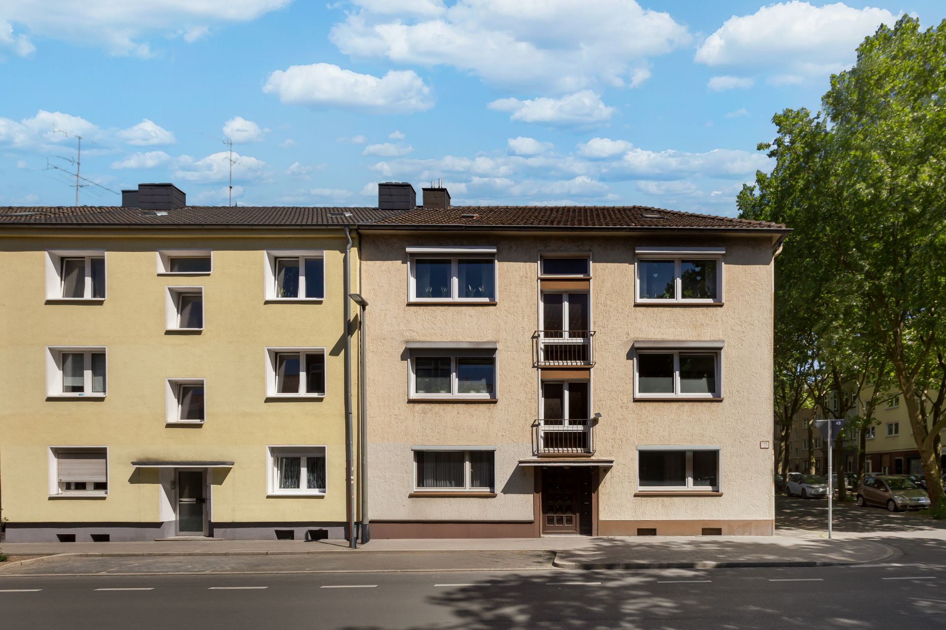 Zwei mehrstöckige Wohnhäuser mit mehreren Fenstern an einer Straße, Bäume im Hintergrund.