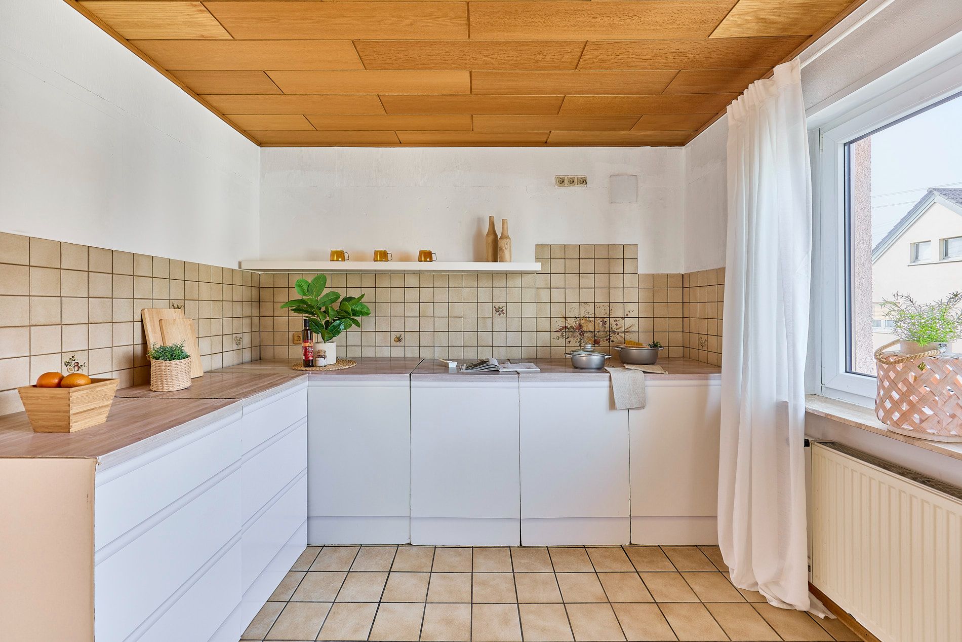 Helle Küche mit weißen Schränken, Holzdach, Fliesenspiegel und großem Fenster mit weißen Vorhängen.