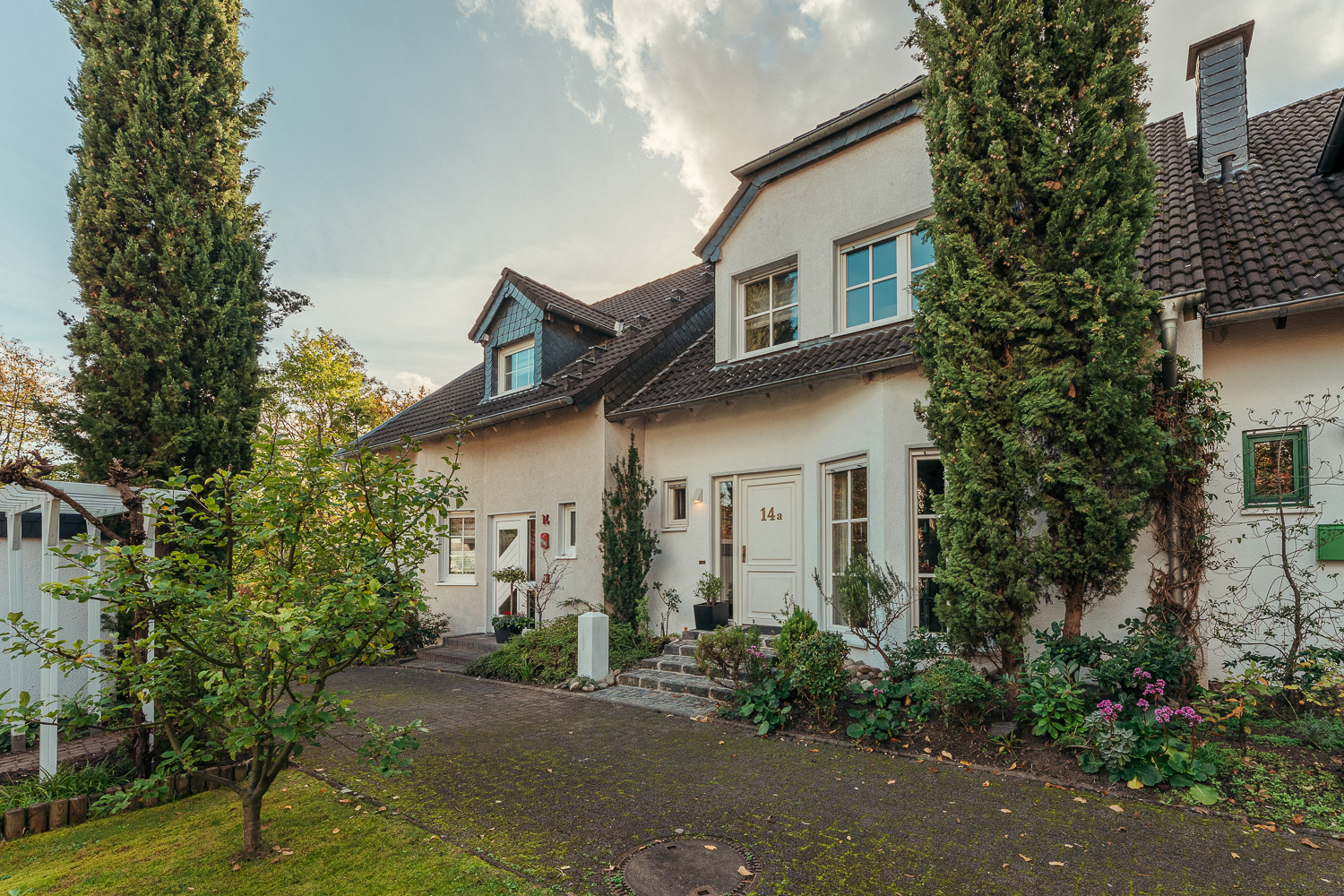 Reihenhaus mit zwei Eingangstüren, Vorgarten, Bäumen und Sträuchern an einem sonnigen Tag.