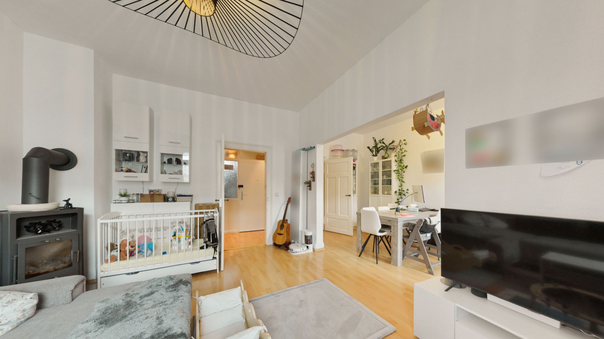Helles Wohnzimmer mit Kinderbett, Sofa, Fernseher und Blick ins Esszimmer mit Esstisch und Stühlen.