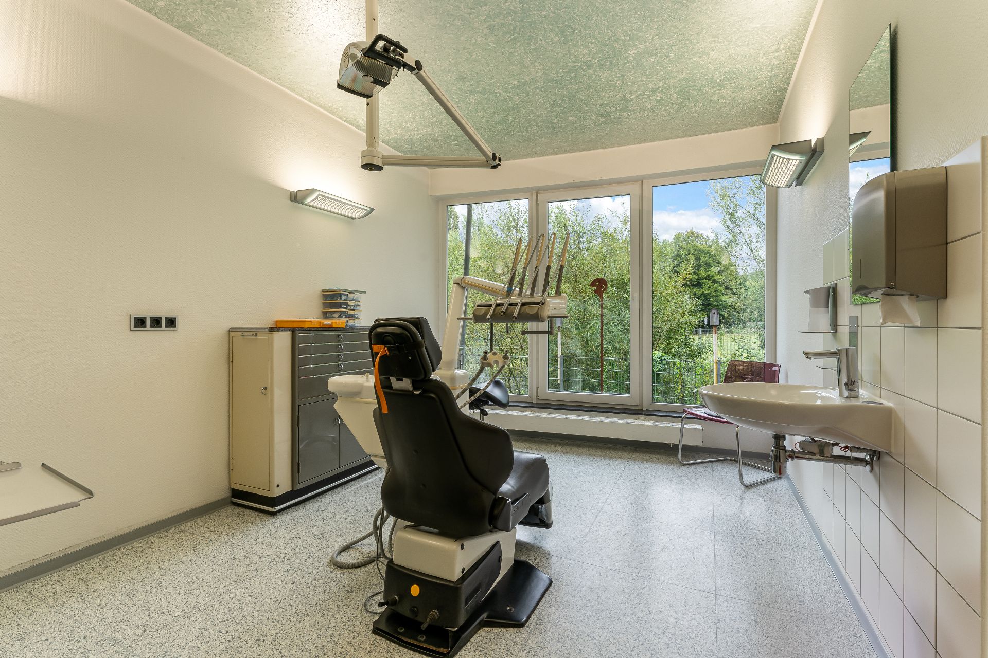 Zahnbehandlungsraum mit Untersuchungsstuhl, Waschbecken und großem Fenster mit Blick ins Grüne.