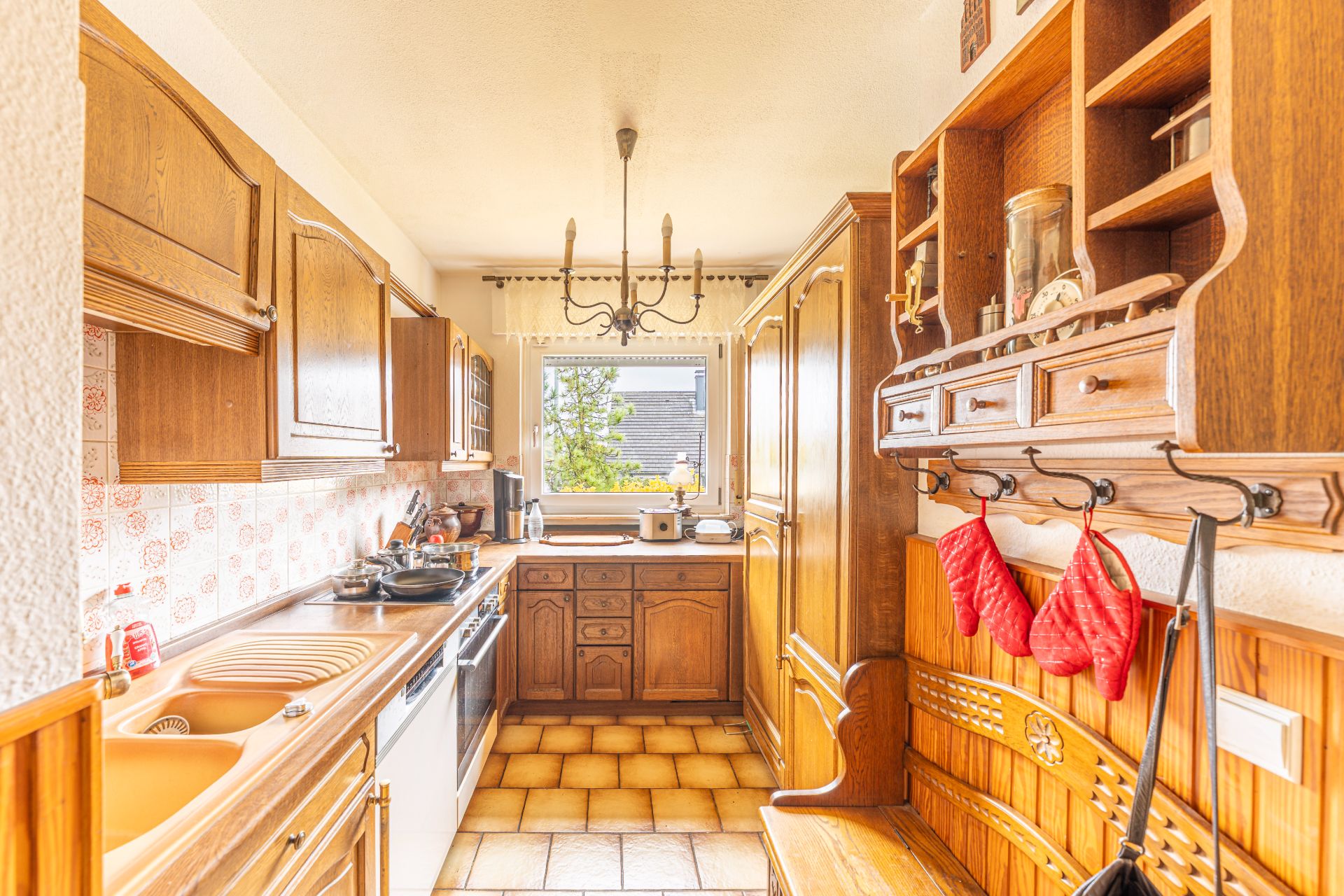 Helle Küche mit Holzschränken, Fenster am Ende und roten Topfhandschuhen an der Wand.