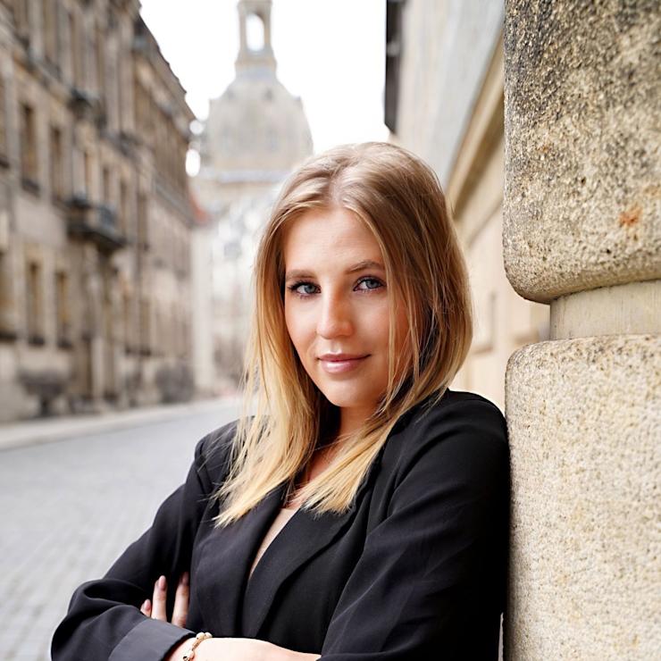 Eine junge Frau mit blondem Haar lehnt an einer Steinmauer in einer Altstadtstraße.