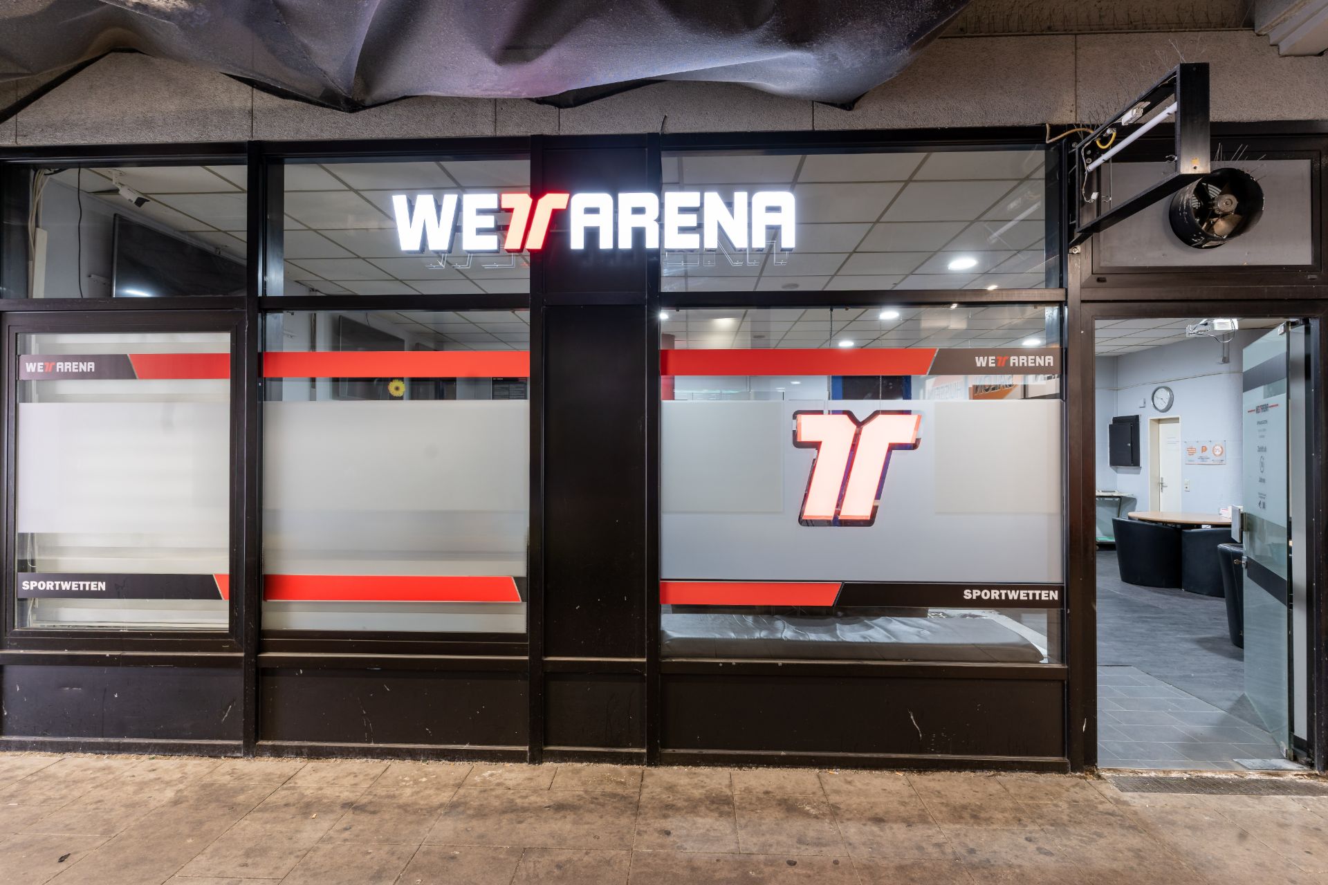 Eingang zu einem Geschäftsraum mit großen Fenstern und beleuchtetem Schild Wetarena.