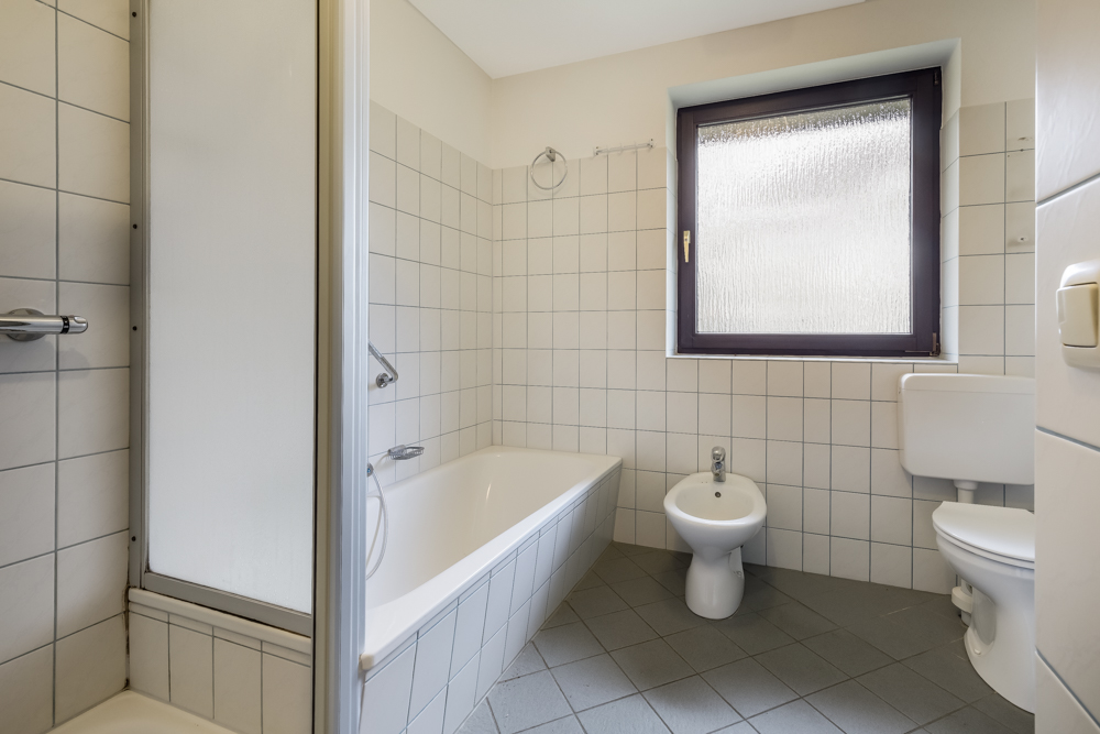 Helles Badezimmer mit Badewanne, Bidet, WC und großem Fenster, weiß gefliest.