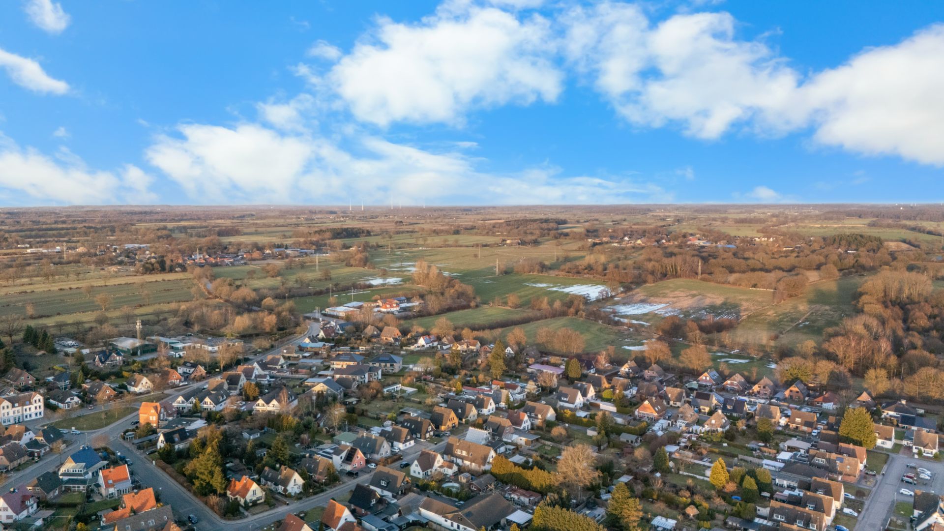 Luftaufnahme eines Wohngebiets mit Häusern, Bäumen und angrenzender Landschaft unter blauem Himmel.