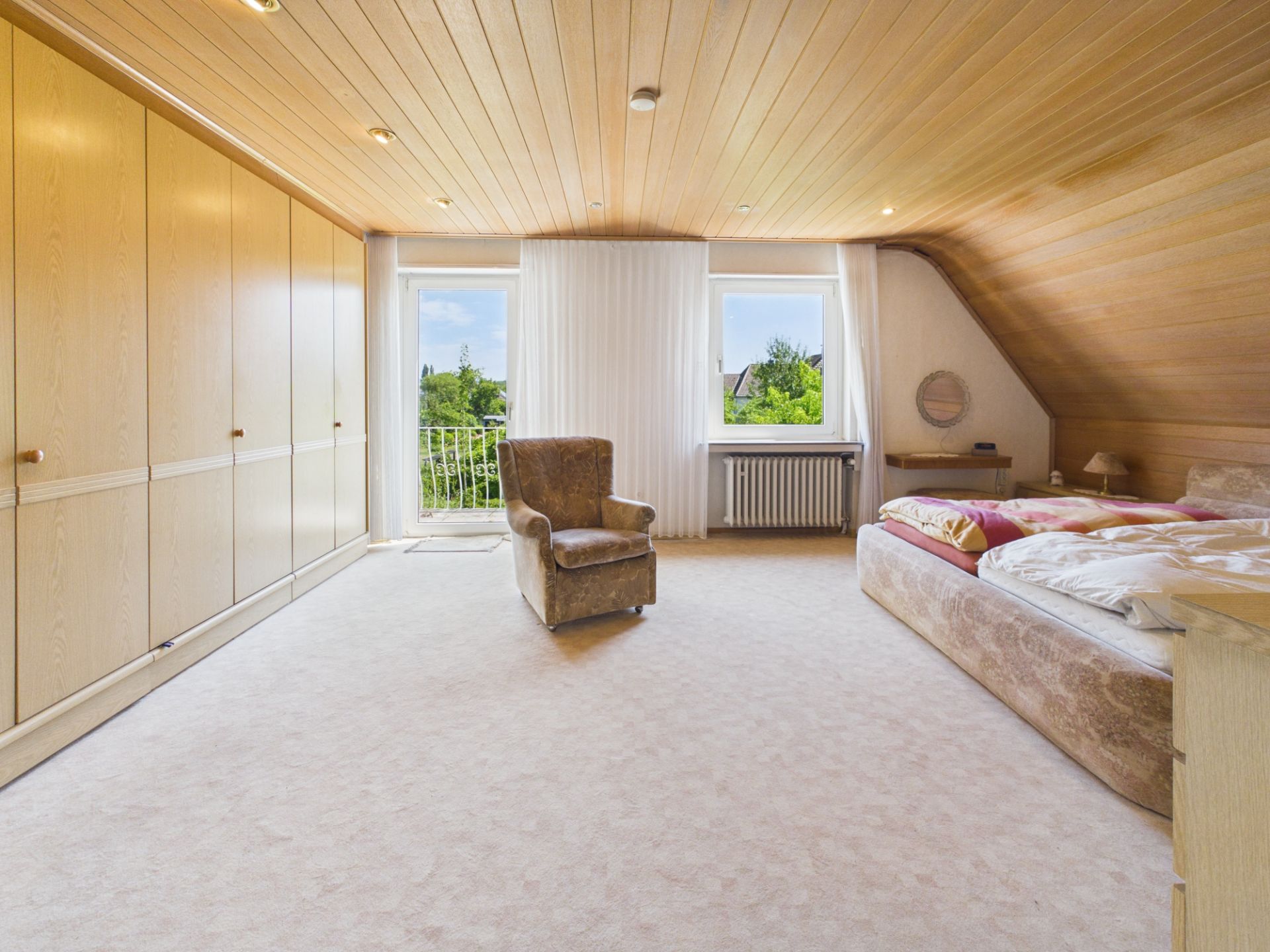 Helles Schlafzimmer mit Doppelbett, Sessel, Einbauschränken und zwei Fenstern mit Blick ins Grüne.