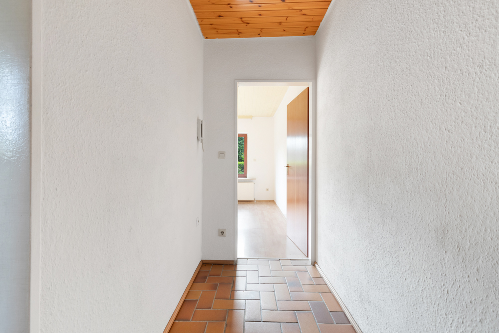 Heller Flur mit braunen Fliesen, weißer Wand und Holztür, Blick in ein Zimmer mit Fenster.
