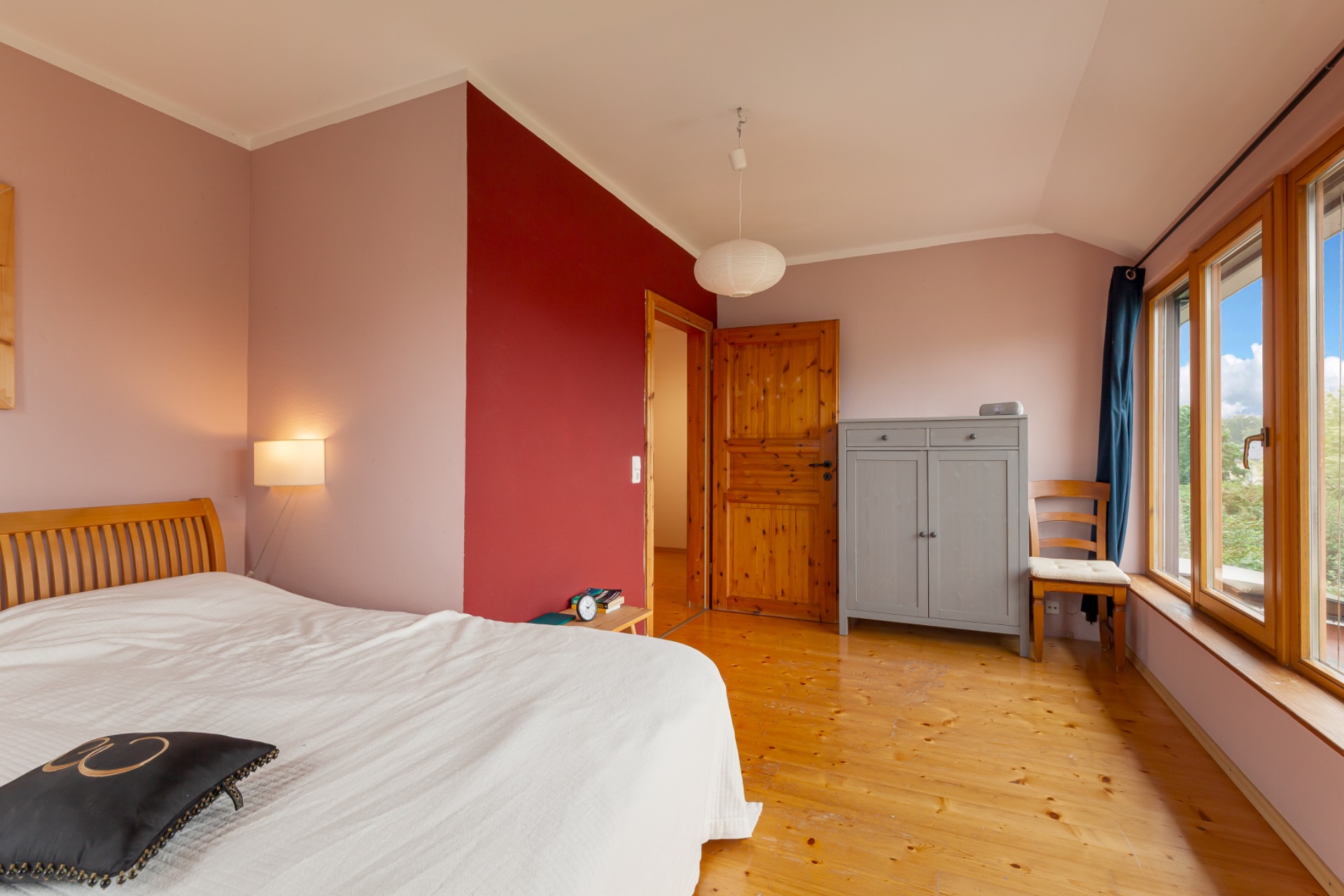 Schlafzimmer mit Holzboden, Bett, grauem Schrank, Stuhl und großem Fenster.