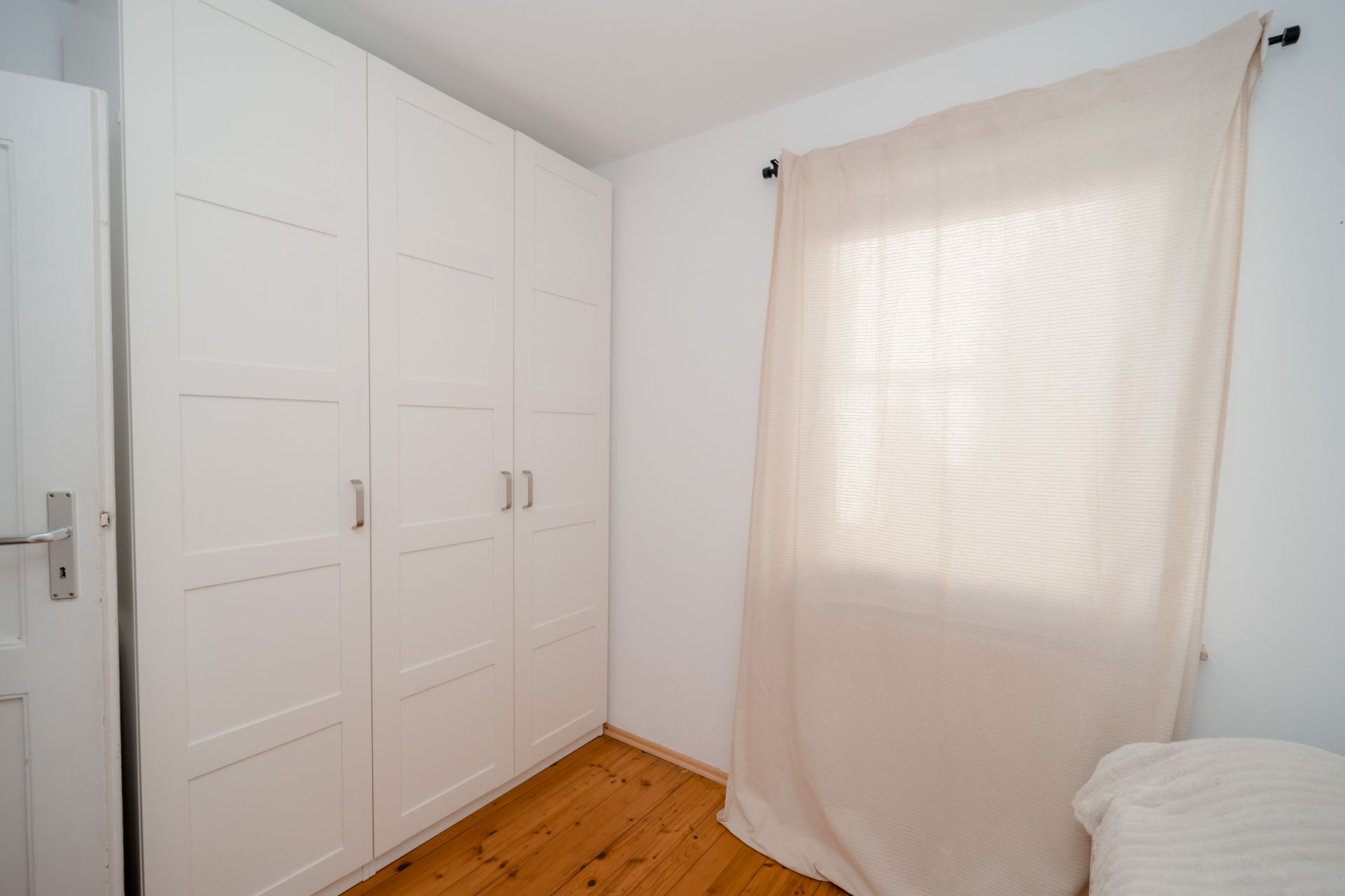 Helles Zimmer mit weißem Kleiderschrank, Holzboden und Fenster mit Vorhang.