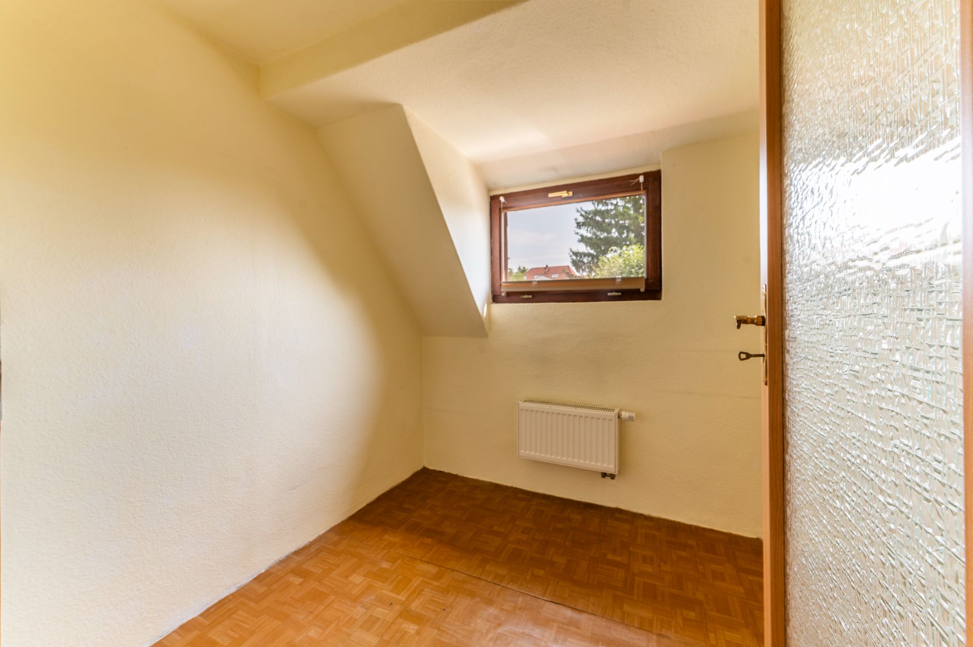 Kleiner leerer Raum mit Parkettboden, Dachschräge, Fenster und Heizkörper.