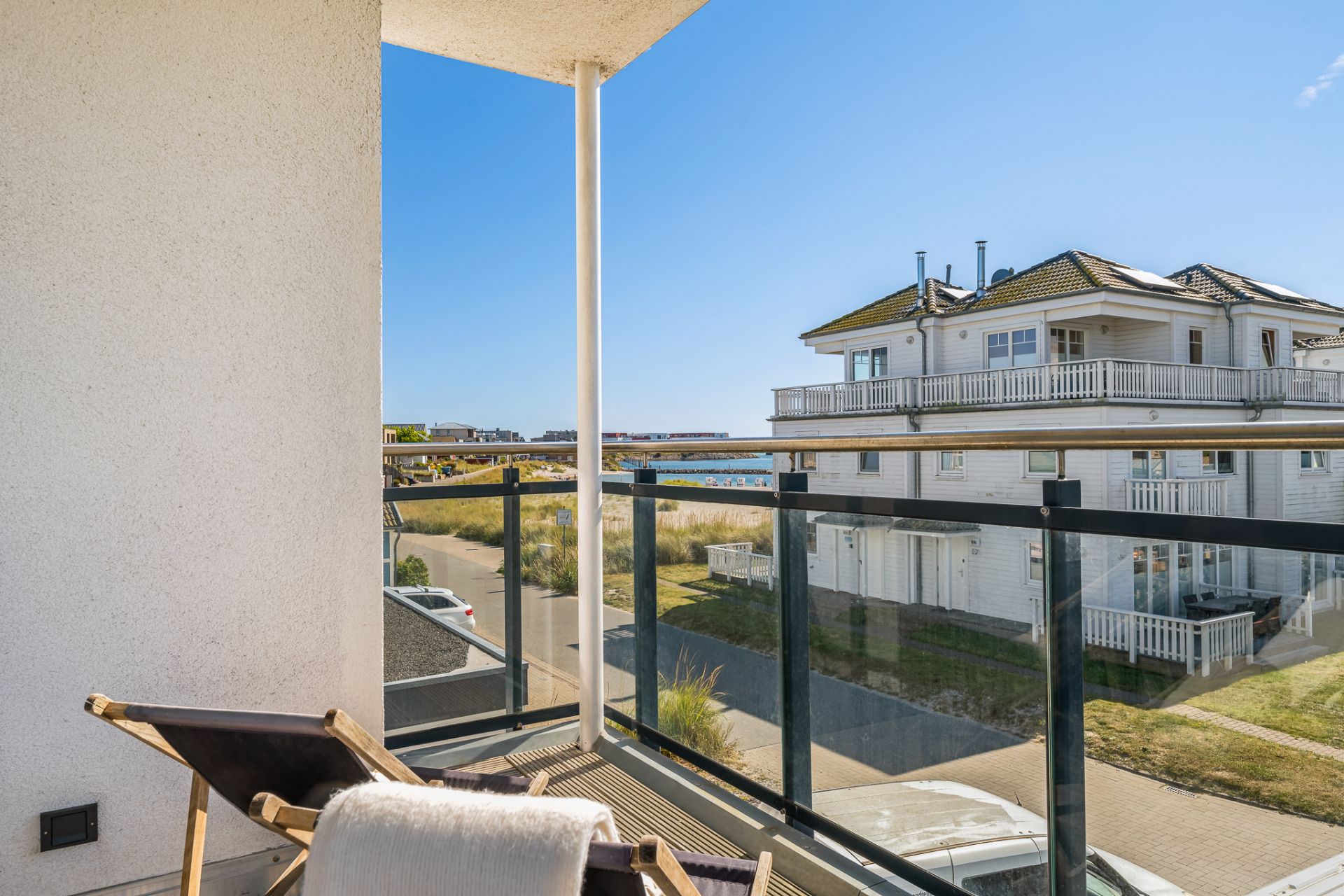 Balkon mit Liegestuhl, Glasgeländer und Blick auf ein Wohnhaus bei sonnigem Wetter.